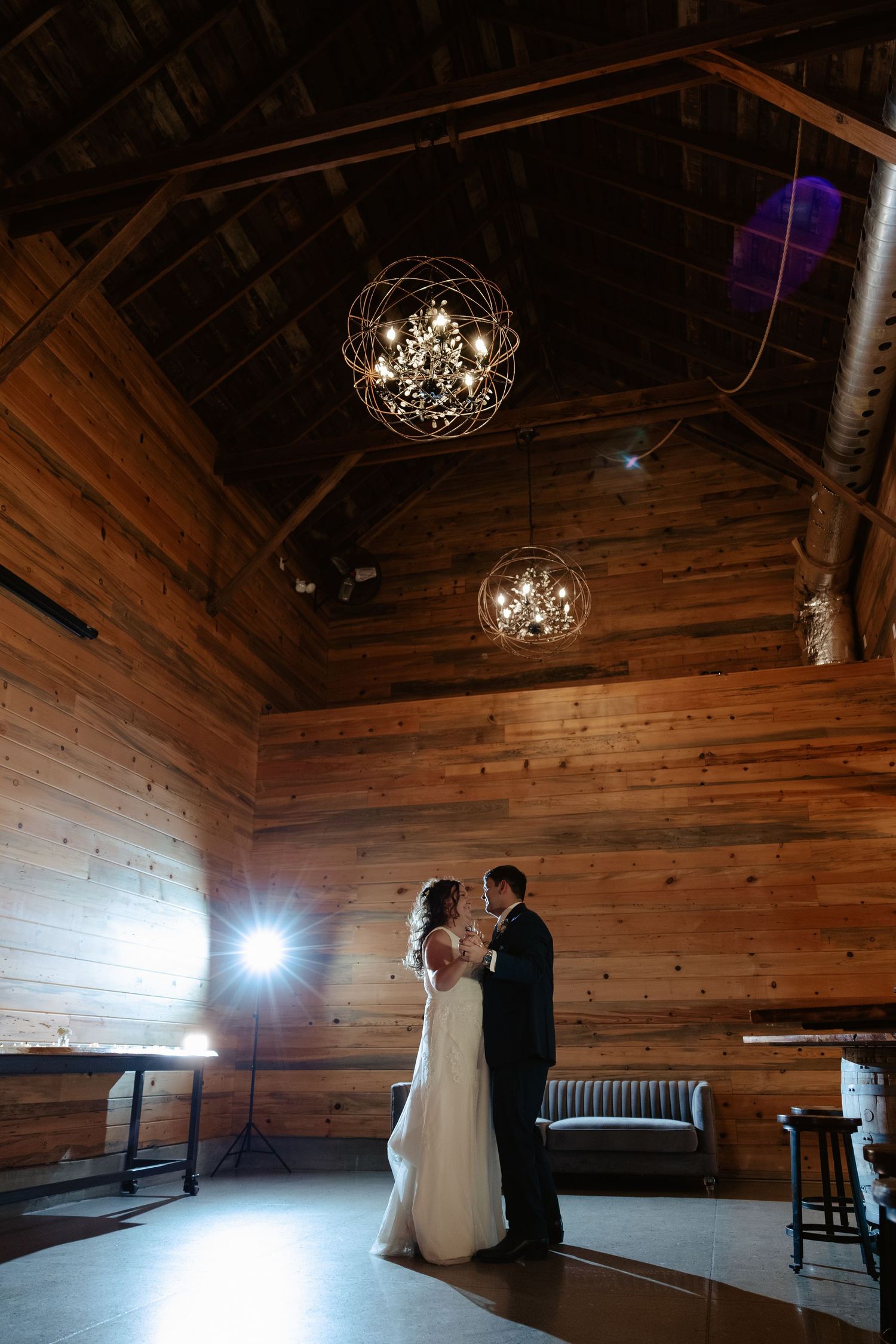 Bride and groom share first dance in rustic wood-paneled barn venue with elegant orb chandeliers overhead at Sparks Barn wedding venue.
