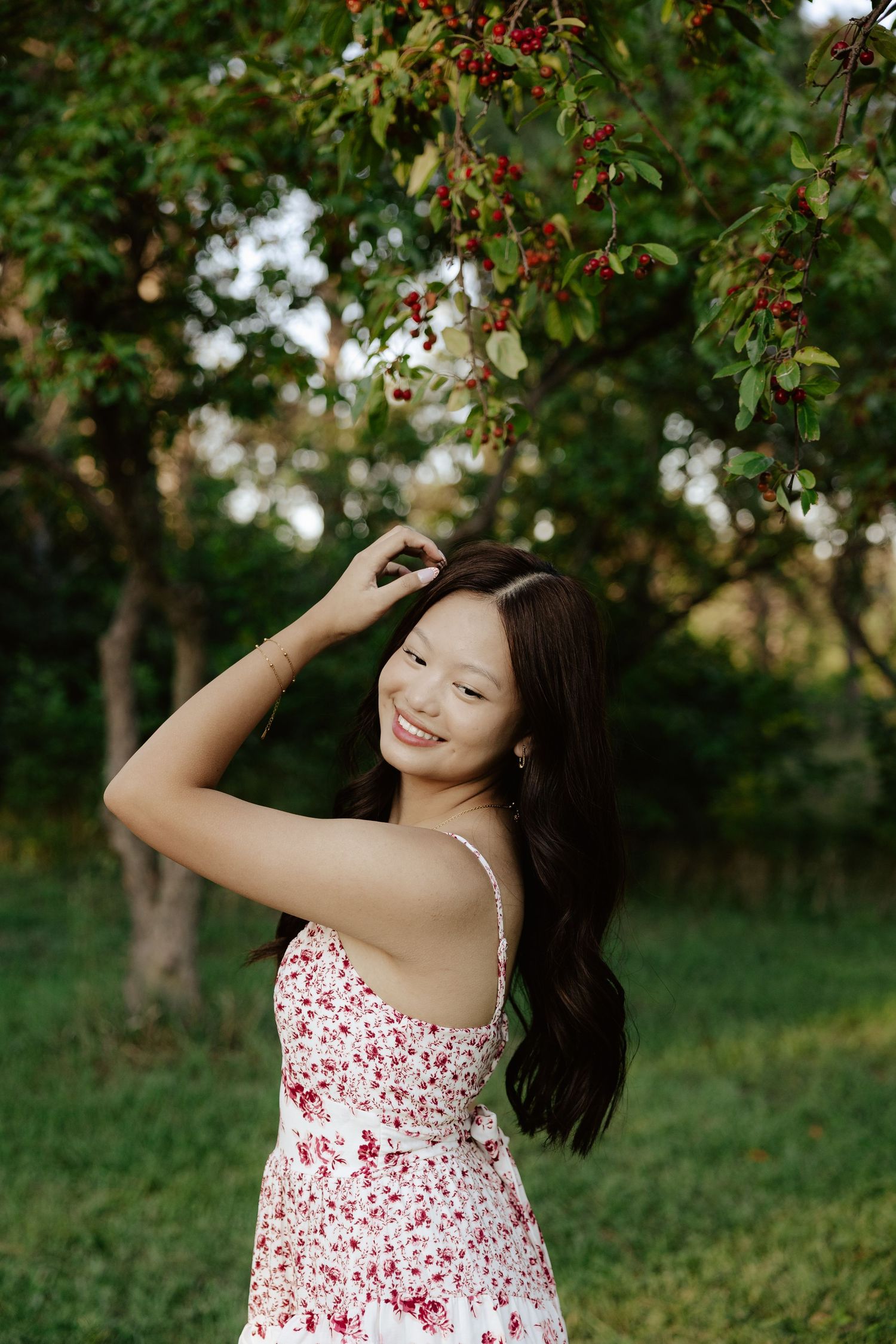 A girl in a white and red floral dress poses outdoors under blooming trees on a sunny day for her senior photos.