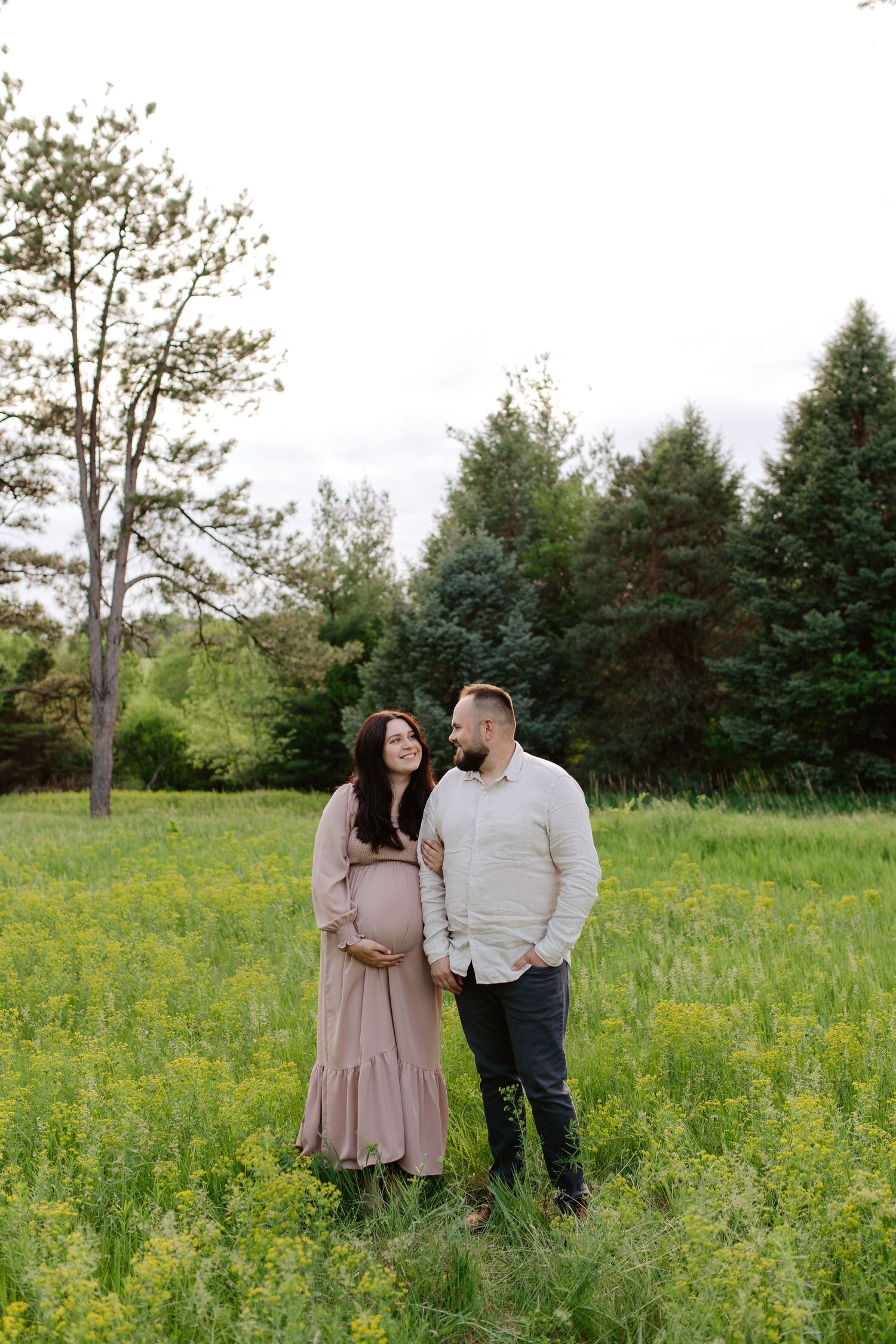 A couple shares a tender moment in a grassy field with pine trees, as they embrace during a maternity photoshoot.
