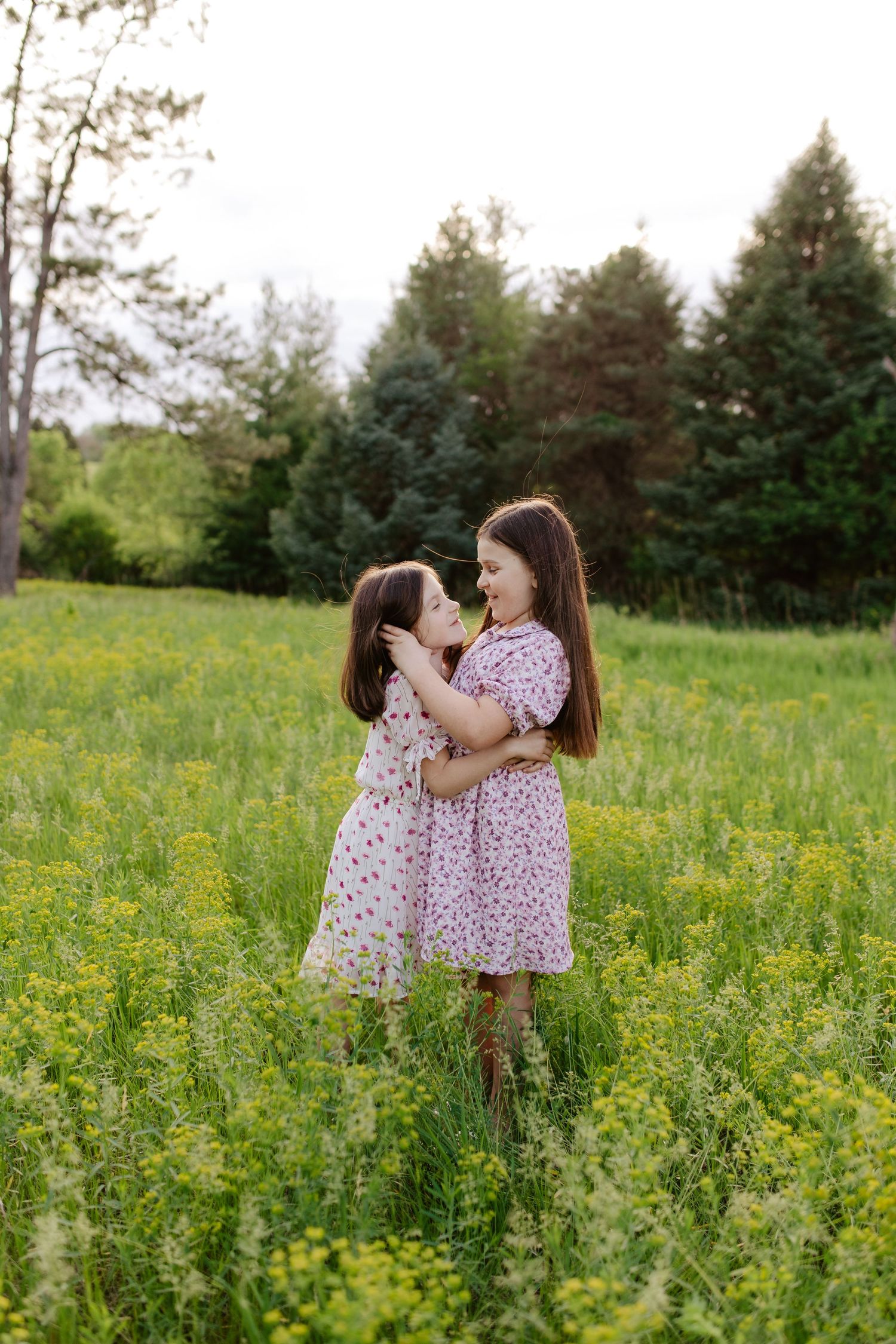 Two children in floral dresses sit together in a grassy meadow with wildflowers on a sunny day.