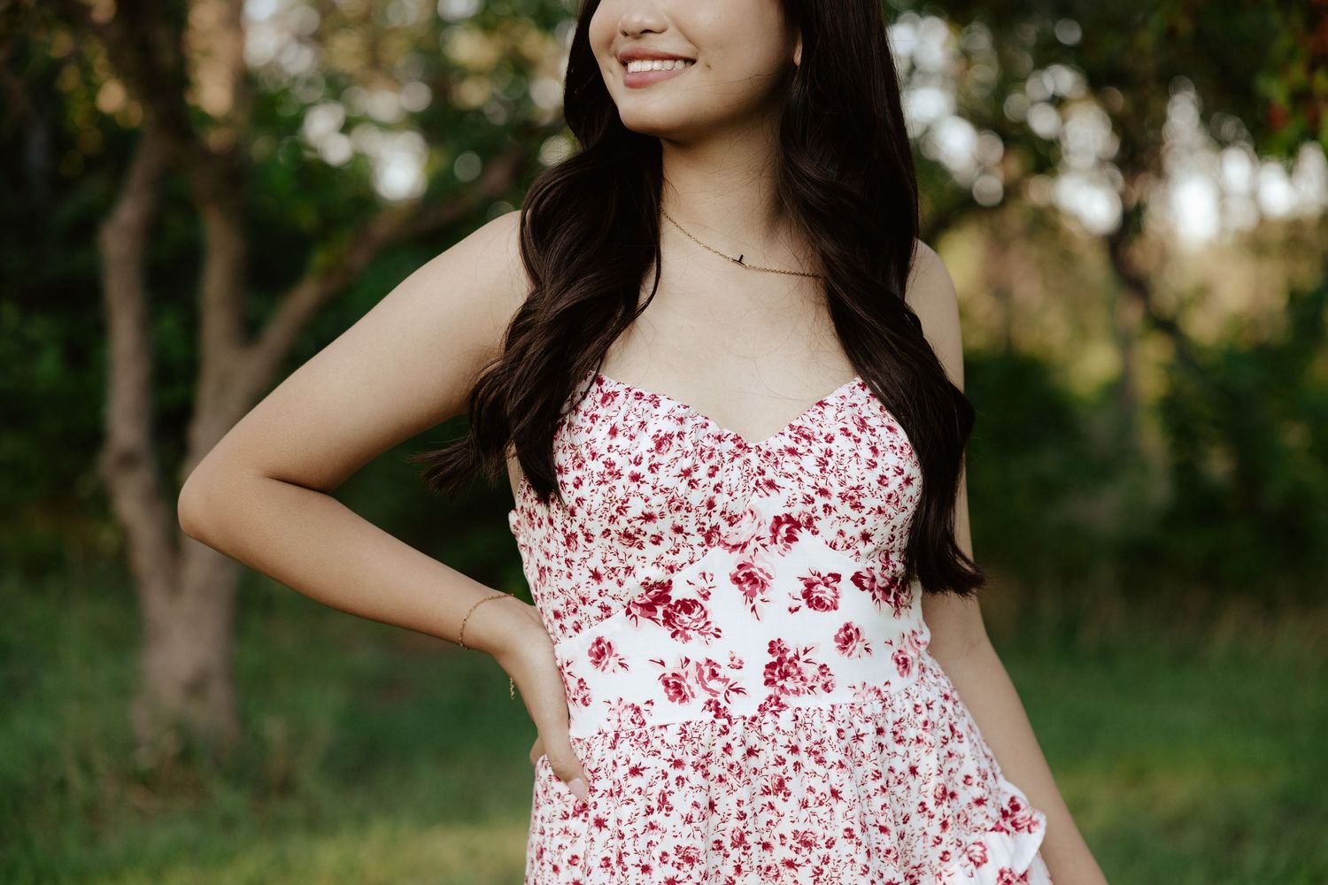 A girl in a white and red floral dress poses outdoors under blooming trees on a sunny day for her senior photos.