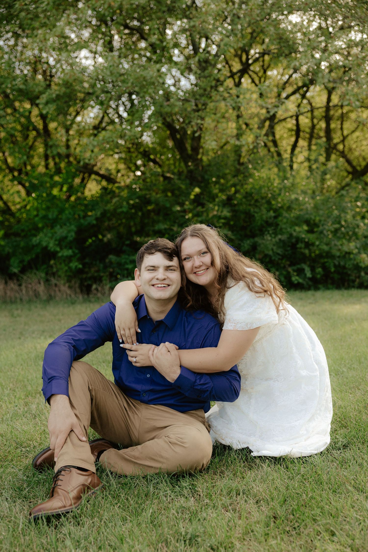 A couple sits together on grass in a natural outdoor setting surrounded by green trees and foliage at Pioneers Park in Lincoln Nebraska.