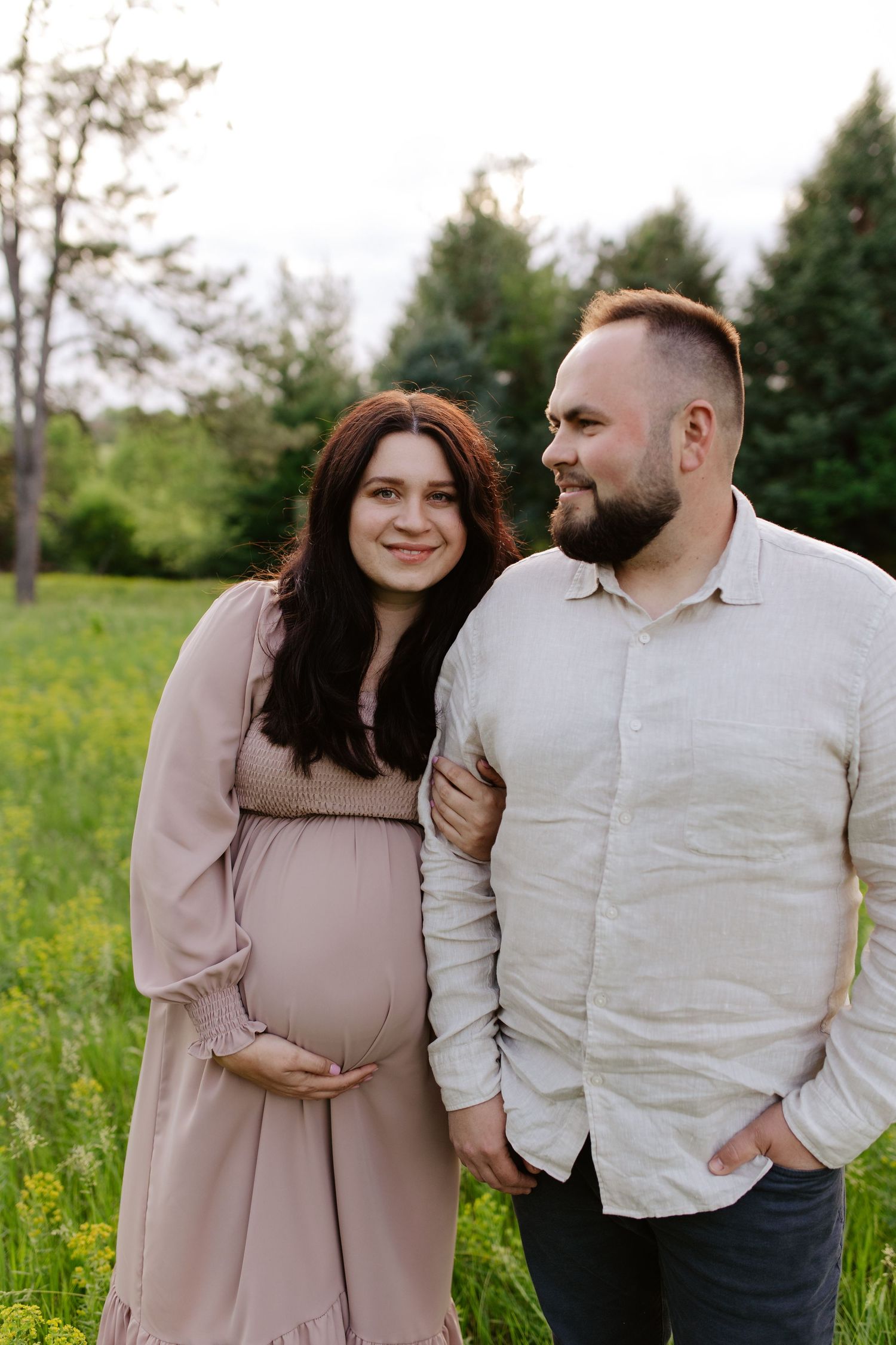 A couple shares a tender moment in a grassy field with pine trees, as they embrace during a maternity photoshoot.