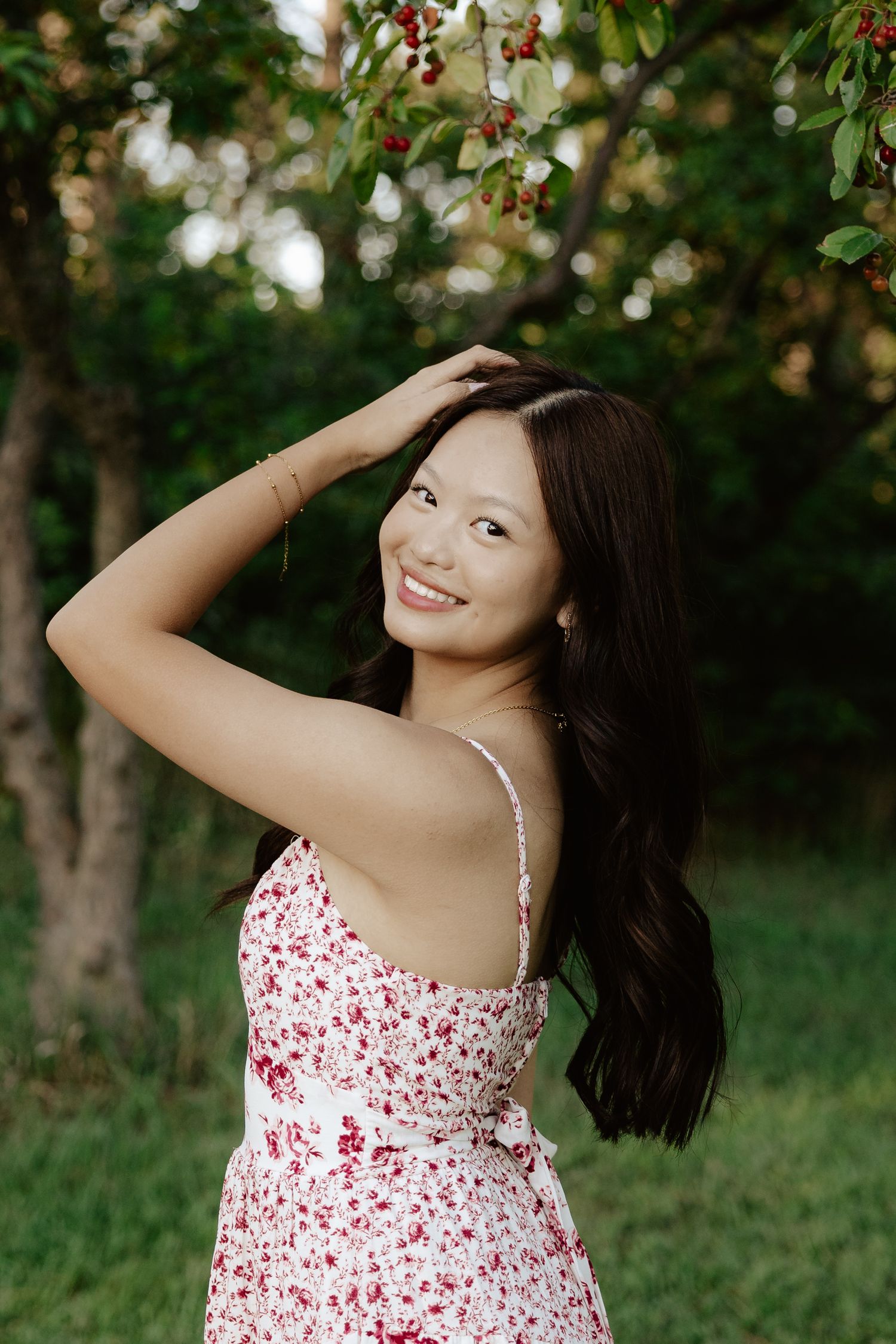 A girl in a white and red floral dress poses outdoors under blooming trees on a sunny day for her senior photos.