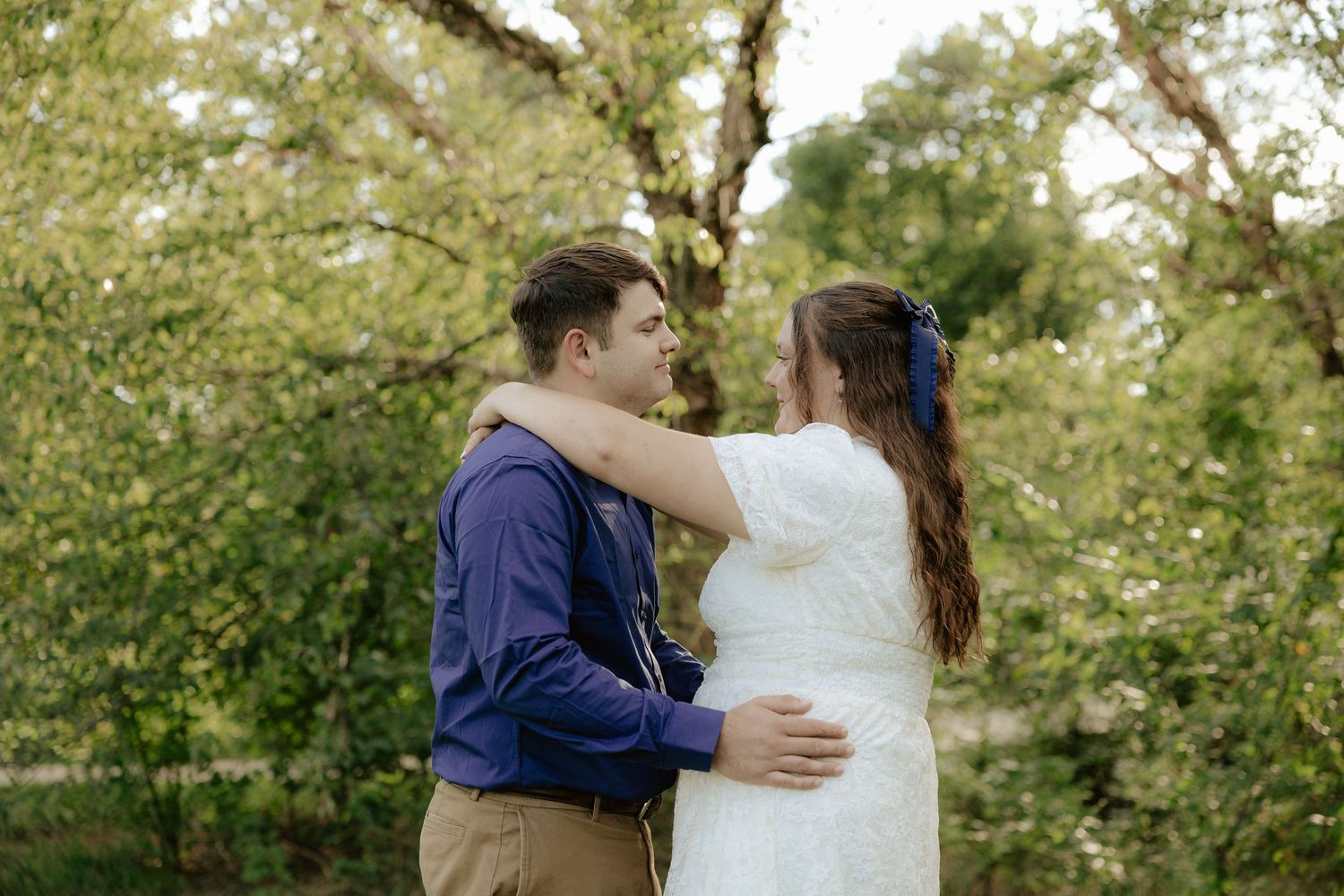 Couple in a romantic embrace standing in a park with lush green foliage in the background.