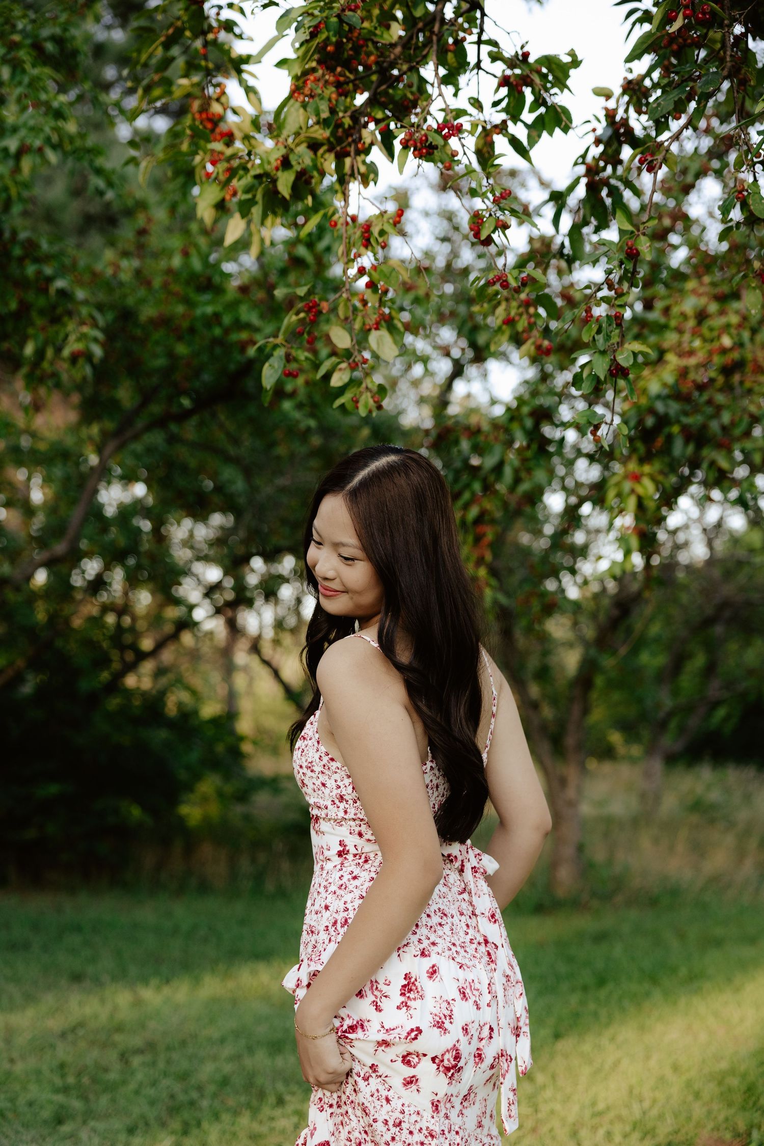 A girl in a white and red floral dress poses outdoors under blooming trees on a sunny day for her senior photos.