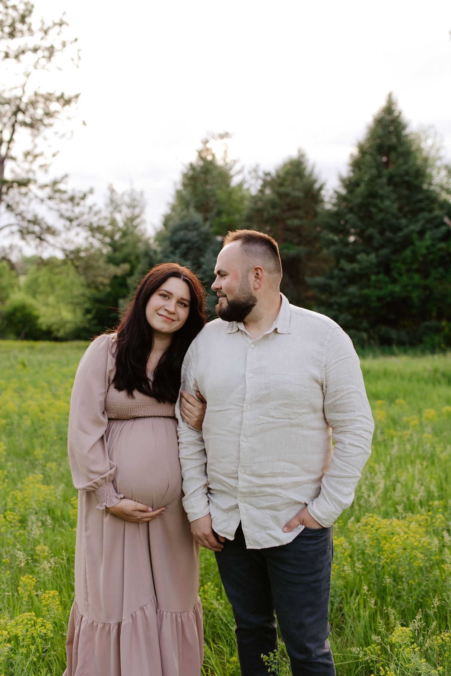 A couple shares a tender moment in a grassy field with pine trees, as they embrace during a maternity photoshoot.