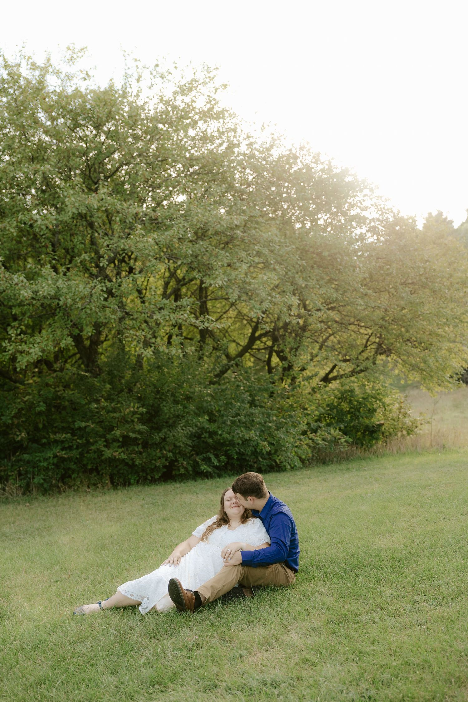 A couple sits together on grass in a natural outdoor setting surrounded by green trees and foliage at Pioneers Park in Lincoln Nebraska.