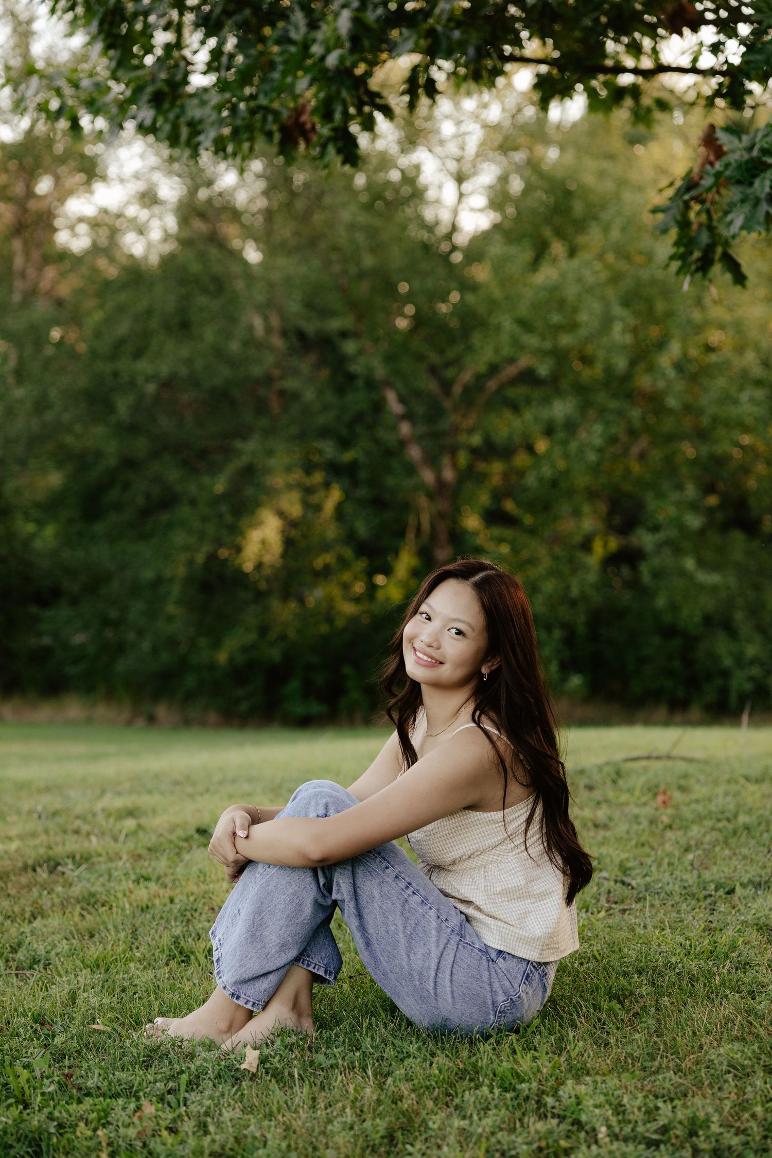 A girl in a beige top and light jeans sits on green grass with trees in the background during golden hour at Pioneers Park in Lincoln NE.