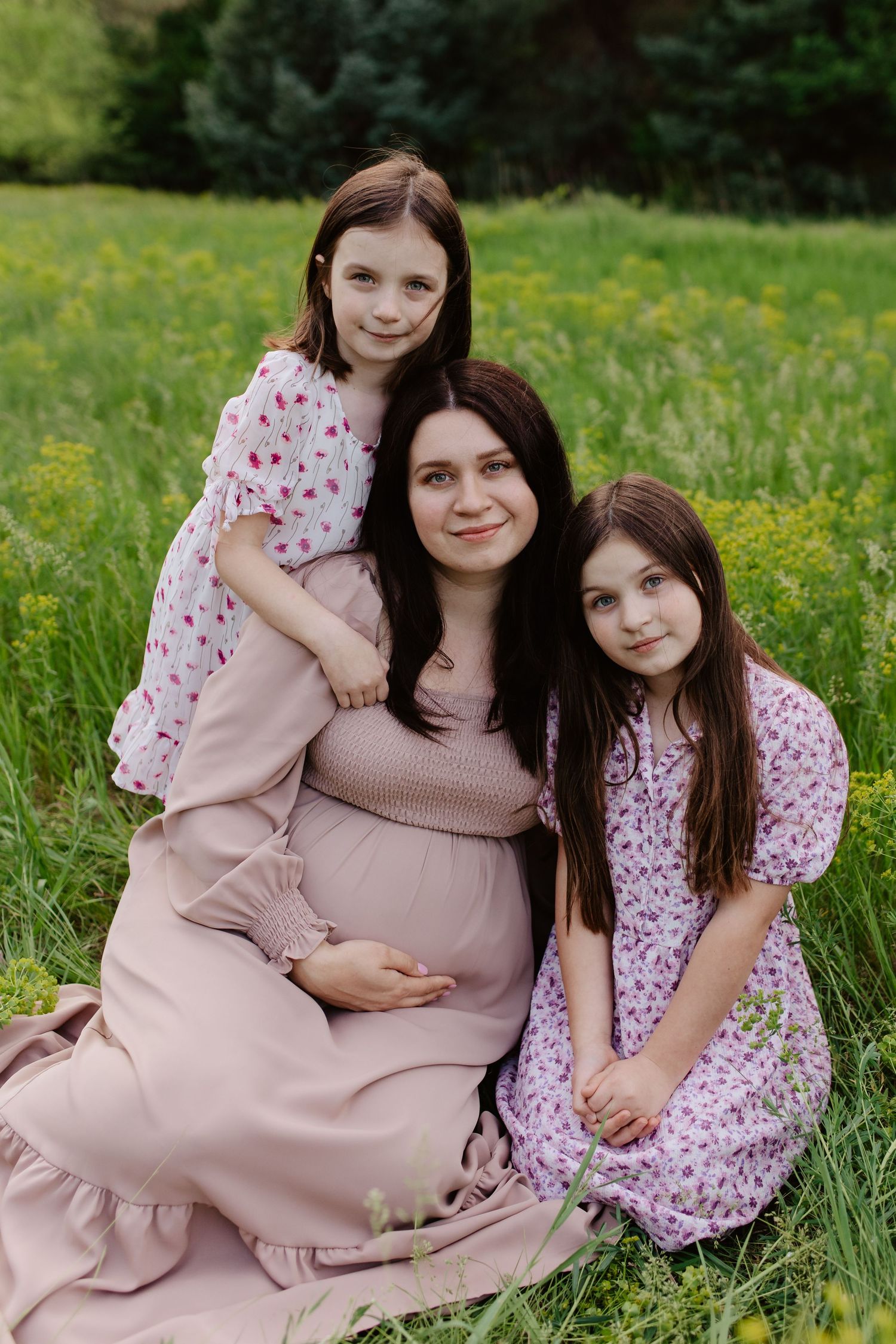 A family wearing pink dresses poses together in a green meadow with yellow wildflowers on a spring day.