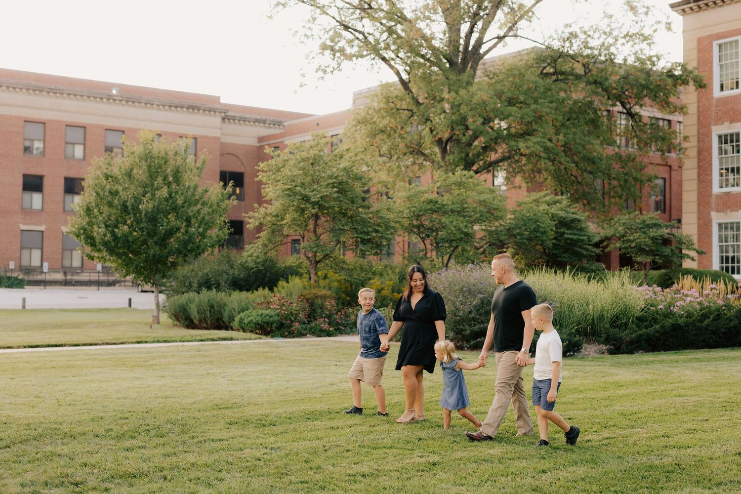 Family walking together on a grassy lawn near brick academic buildings with trees on a sunny day. Downtown UNL family photos