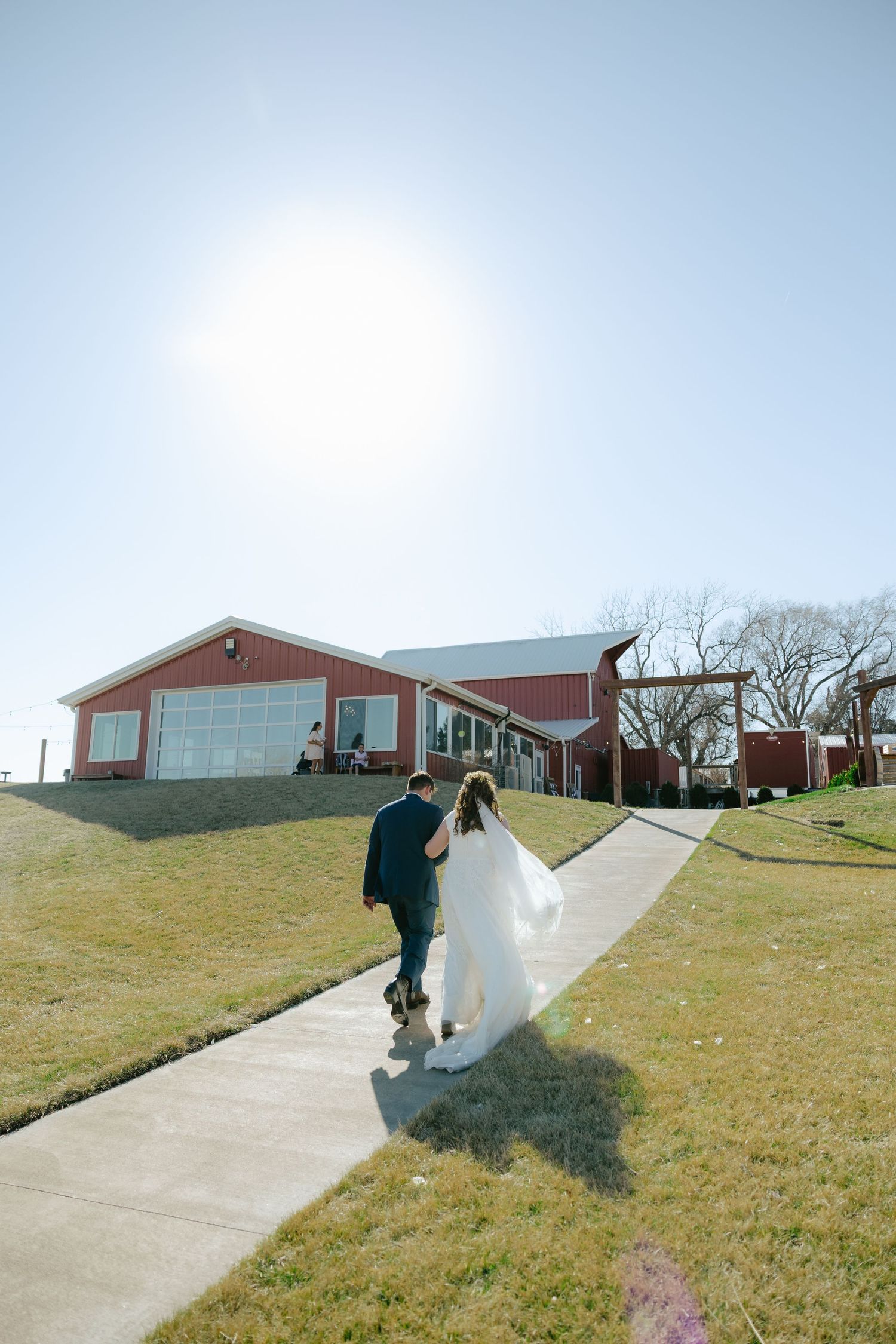 Bride and groom walking hand in hand toward a red barn wedding venue on a sunny day, Sparks Barn in Lousville Nebraska.
