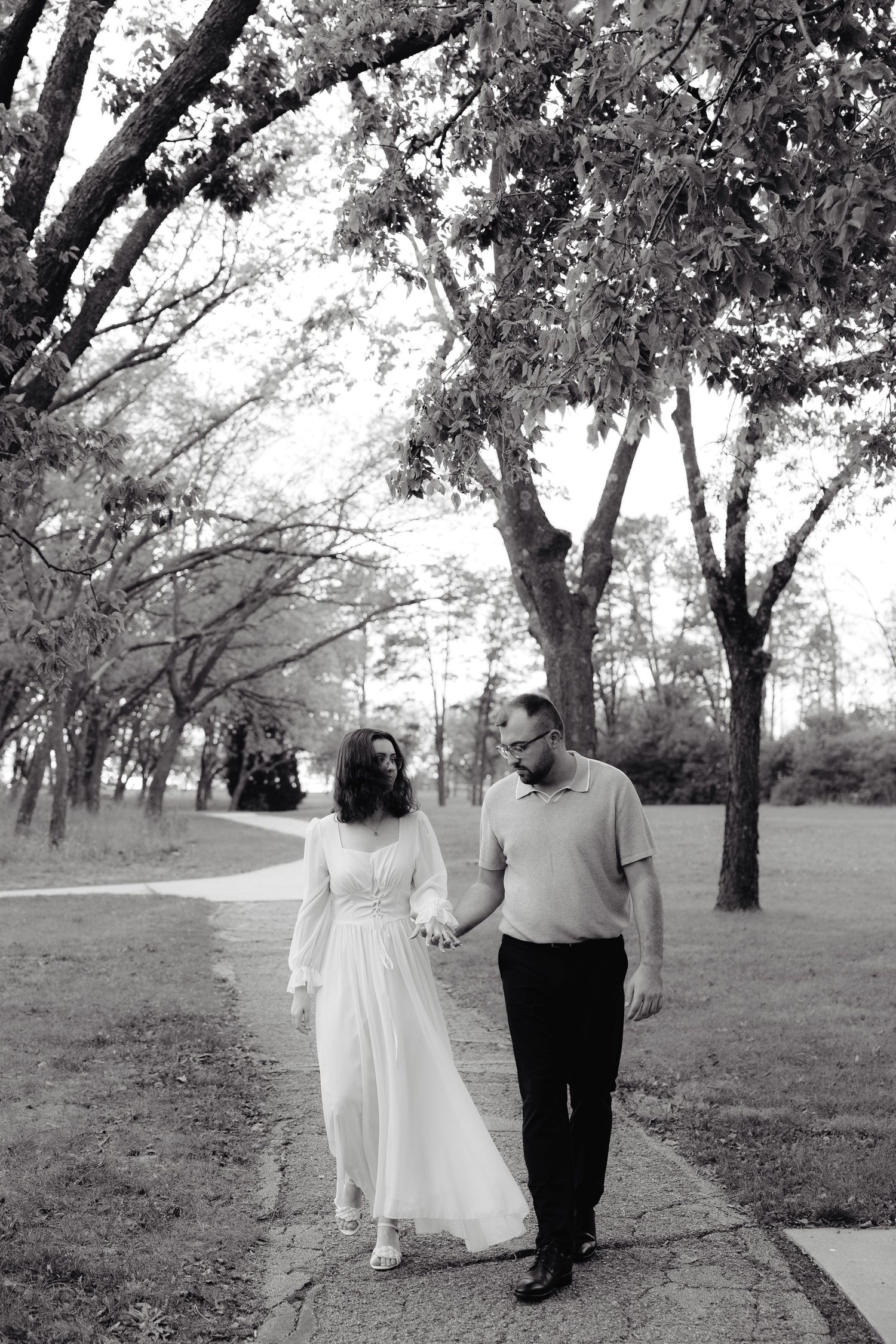Black and white photo of a couple walking together on a tree-lined path in a romantic outdoor setting.