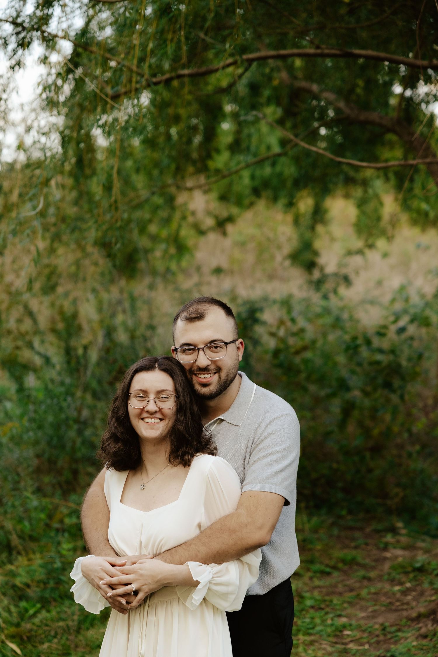A couple embraces in a romantic outdoor photoshoot under green trees and natural lighting.