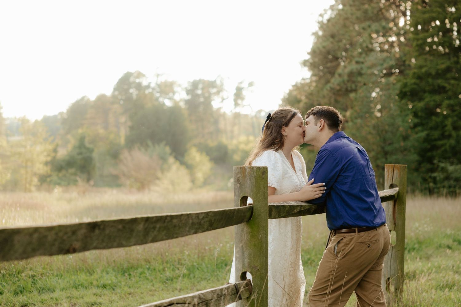 A couple embraces tenderly in an outdoor setting while wearing blue and white outfits in golden sunlight.
