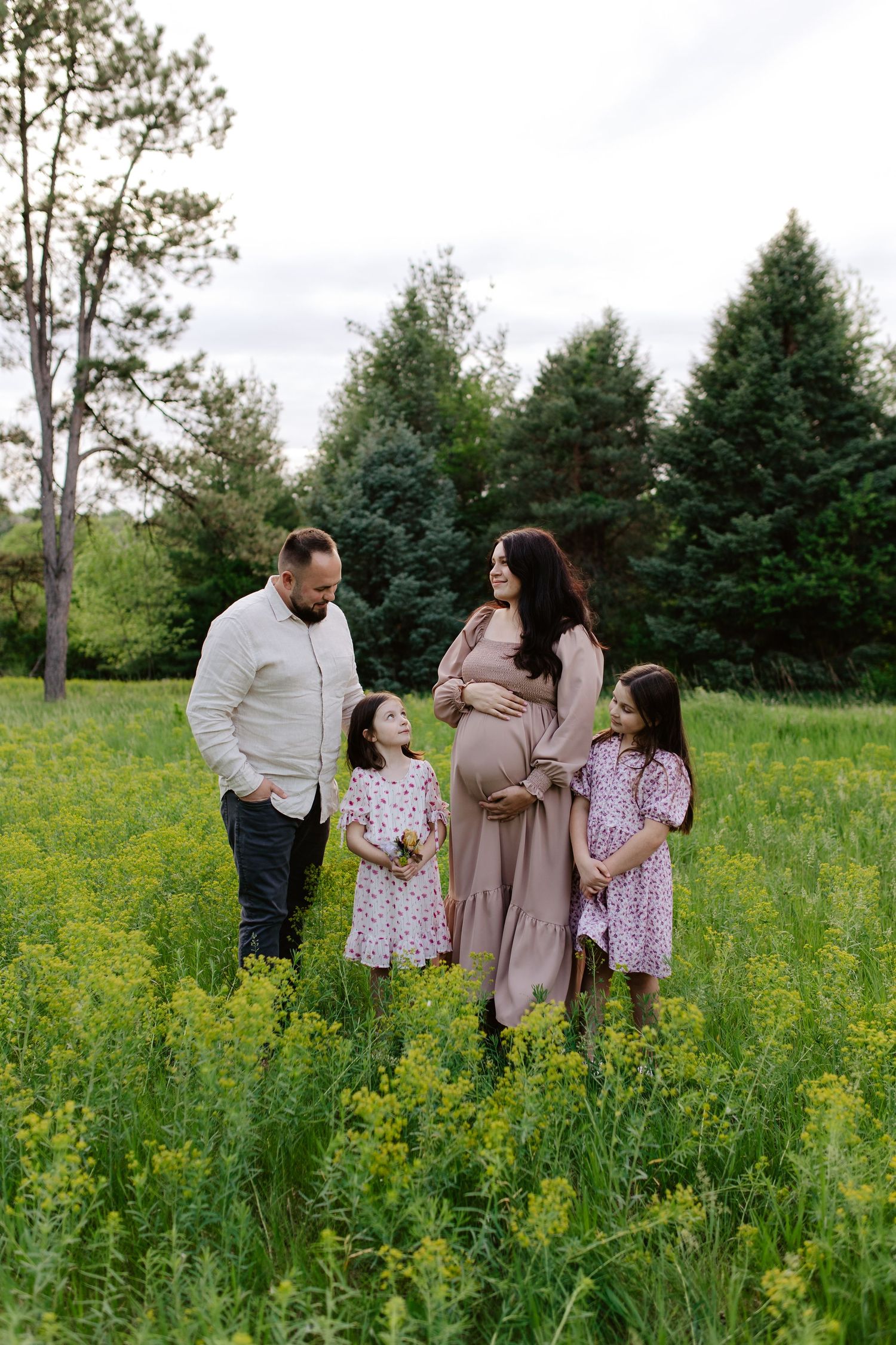 Family stands together in a field of yellow wildflowers with evergreen trees in the background on a cloudy day.