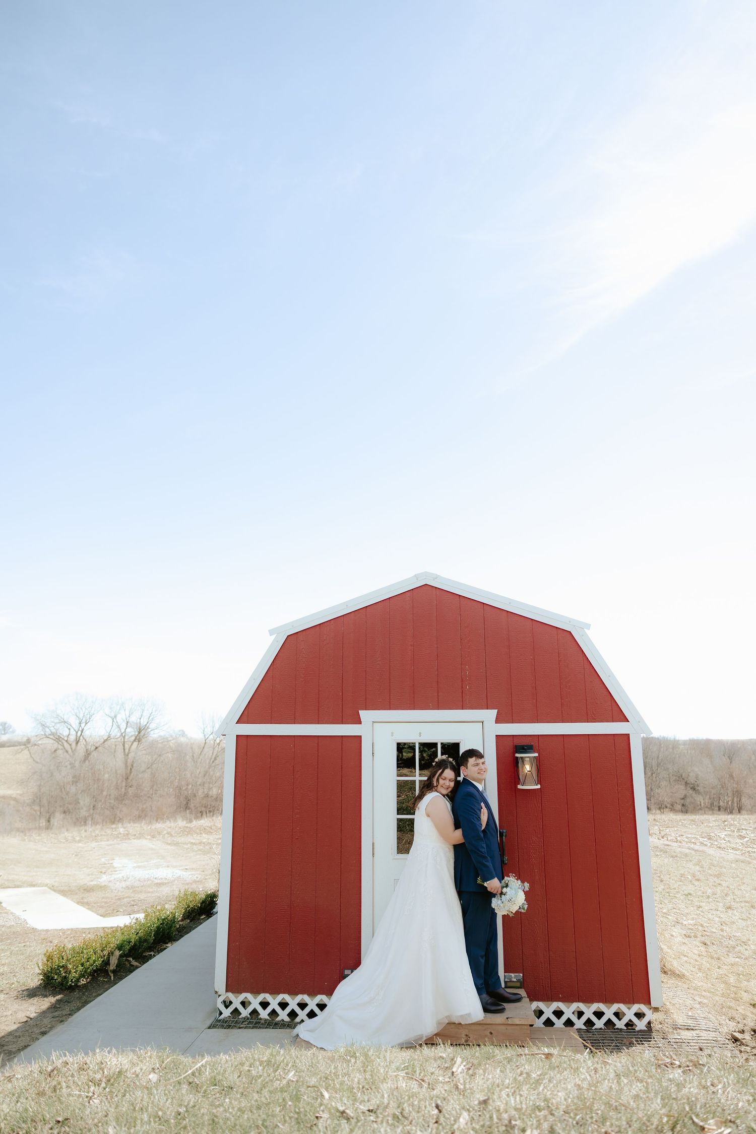 A bride and groom pose together in front of a charming small red barn on their rustic outdoor wedding day.