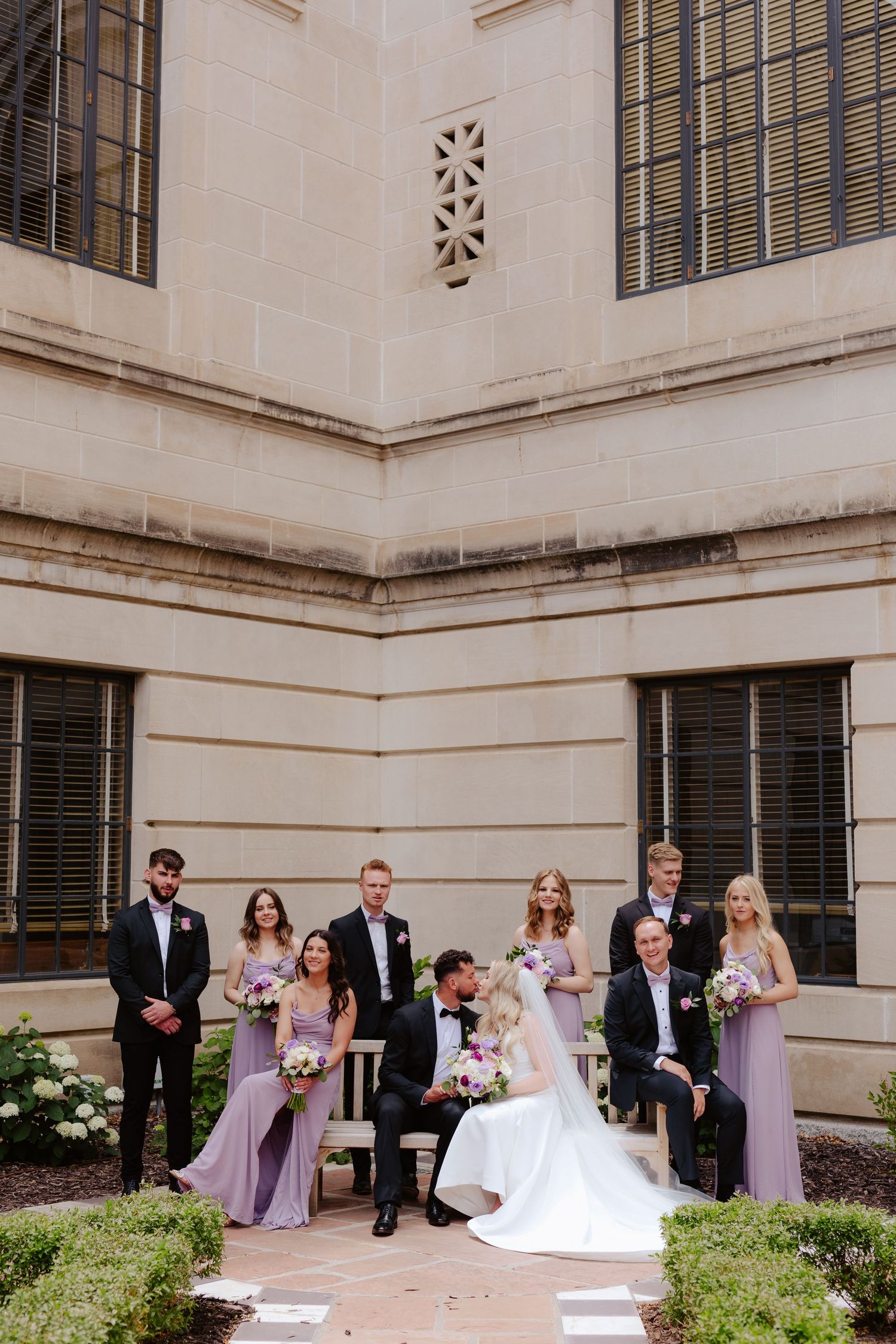 Wedding party poses in an outdoor courtyard with white walls and black shuttered windows at the Nebraska State Capitol building in Lincoln.
