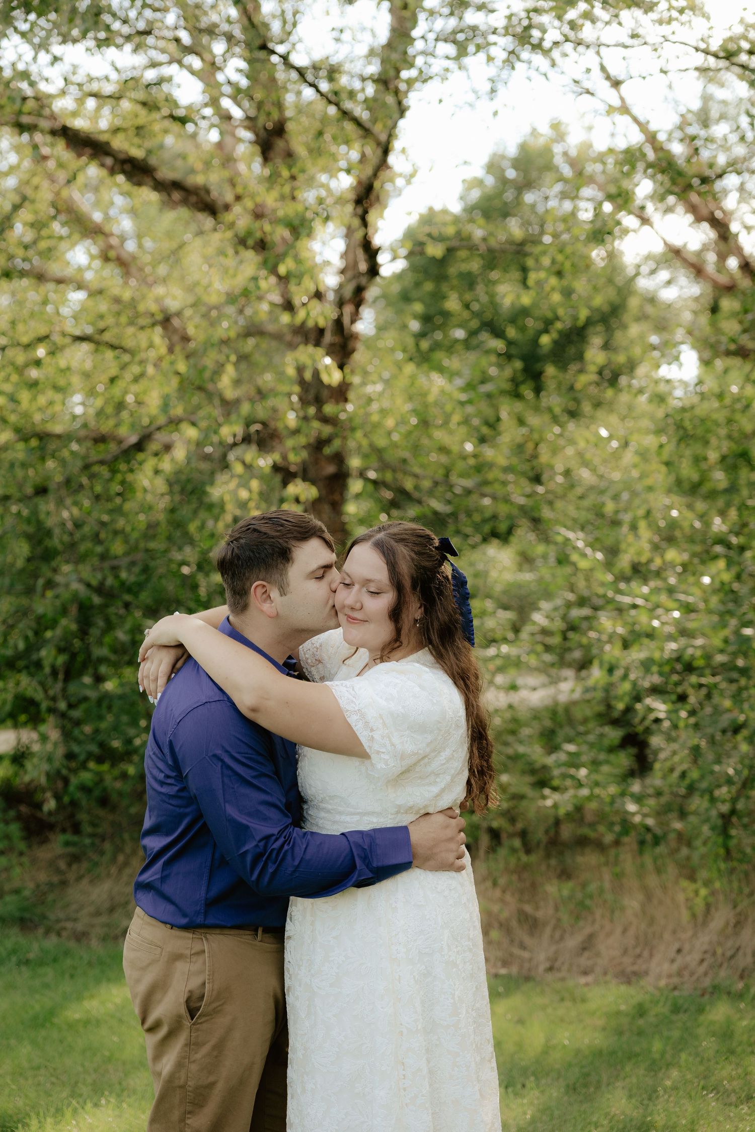 A couple embraces in a romantic outdoor setting among green trees on a sunny day.