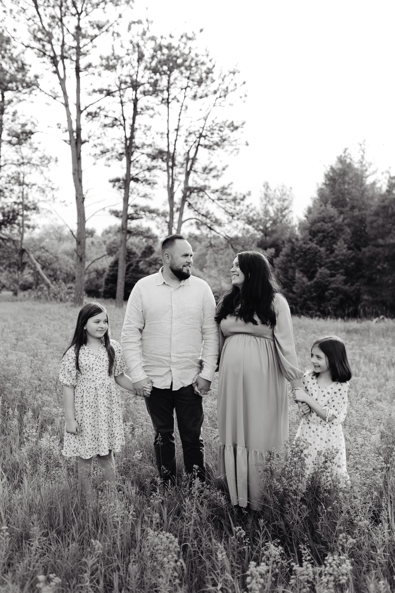 A family walks together holding hands in a green meadow with tall trees in the background on a summer day, maternity photoshoot.