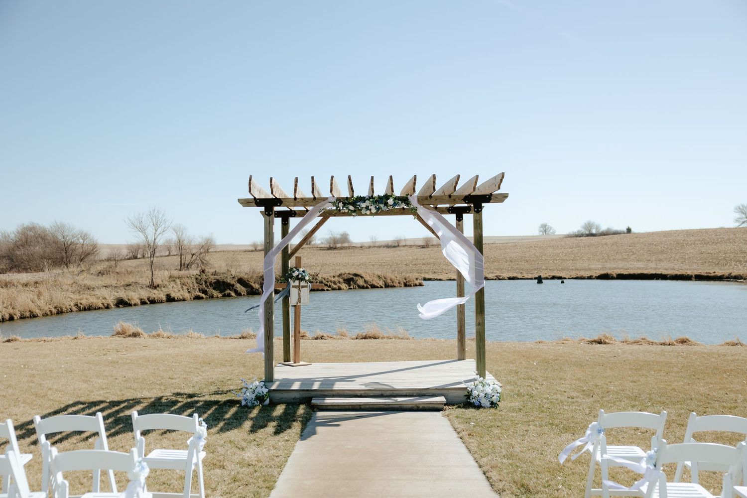 Outdoor wedding altar with wooden cross, white flowing fabric, and blue floral decor beside a serene pond at Sparks Barn in Nebraska