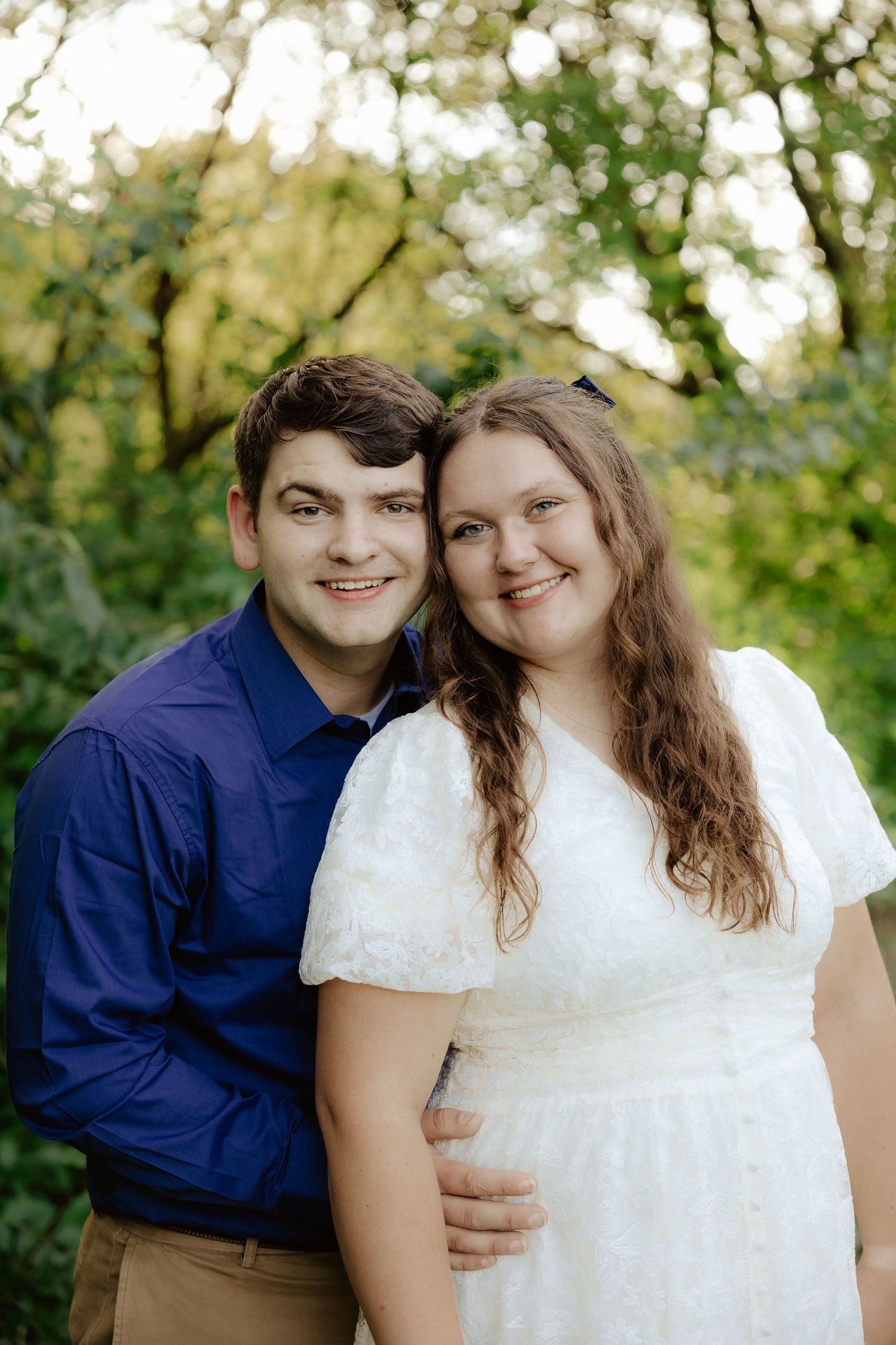 A couple poses together outdoors in wedding attire with white dress and navy blue shirt among green foliage.