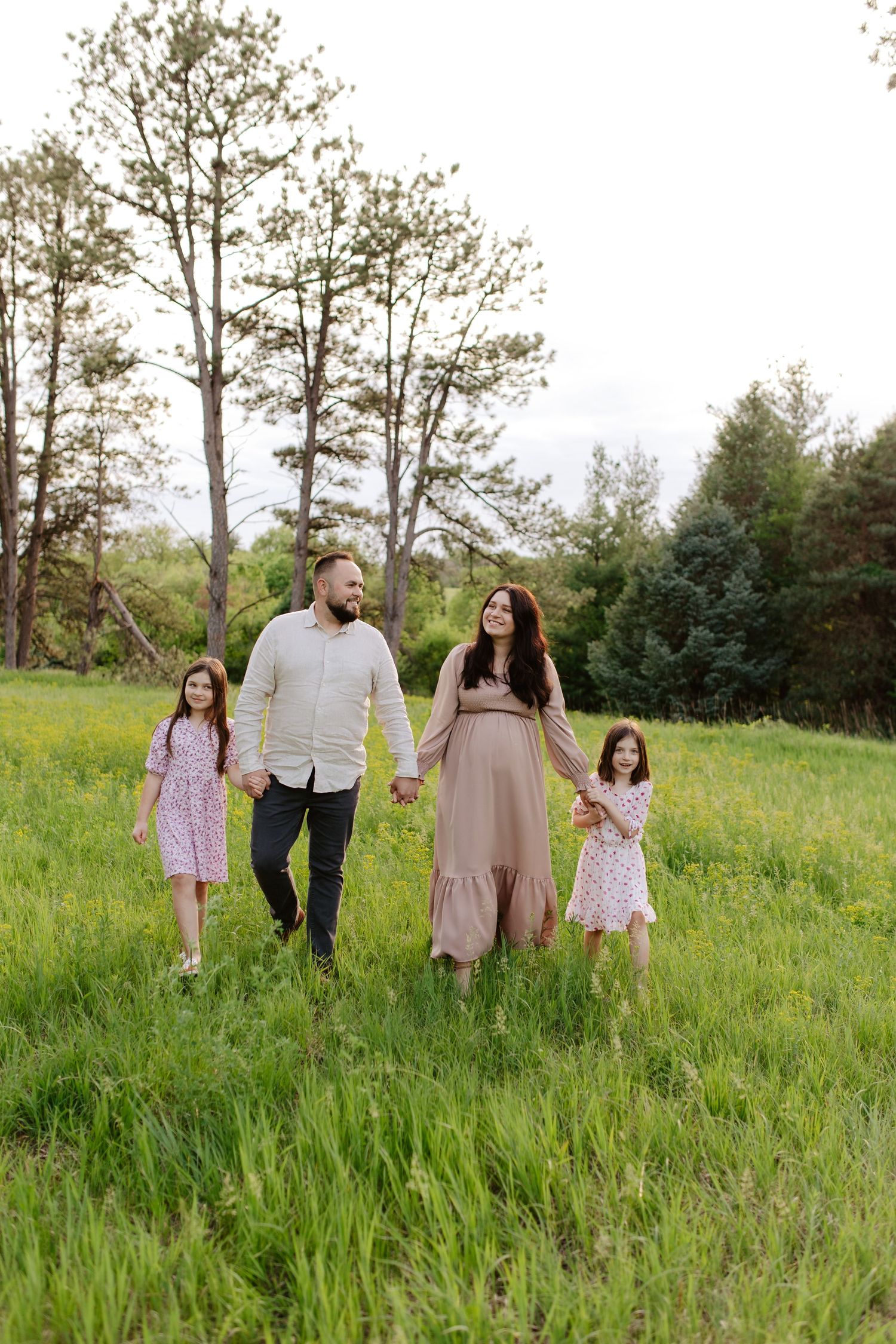 A family walks together holding hands in a green meadow with tall trees in the background on a summer day, maternity photoshoot.