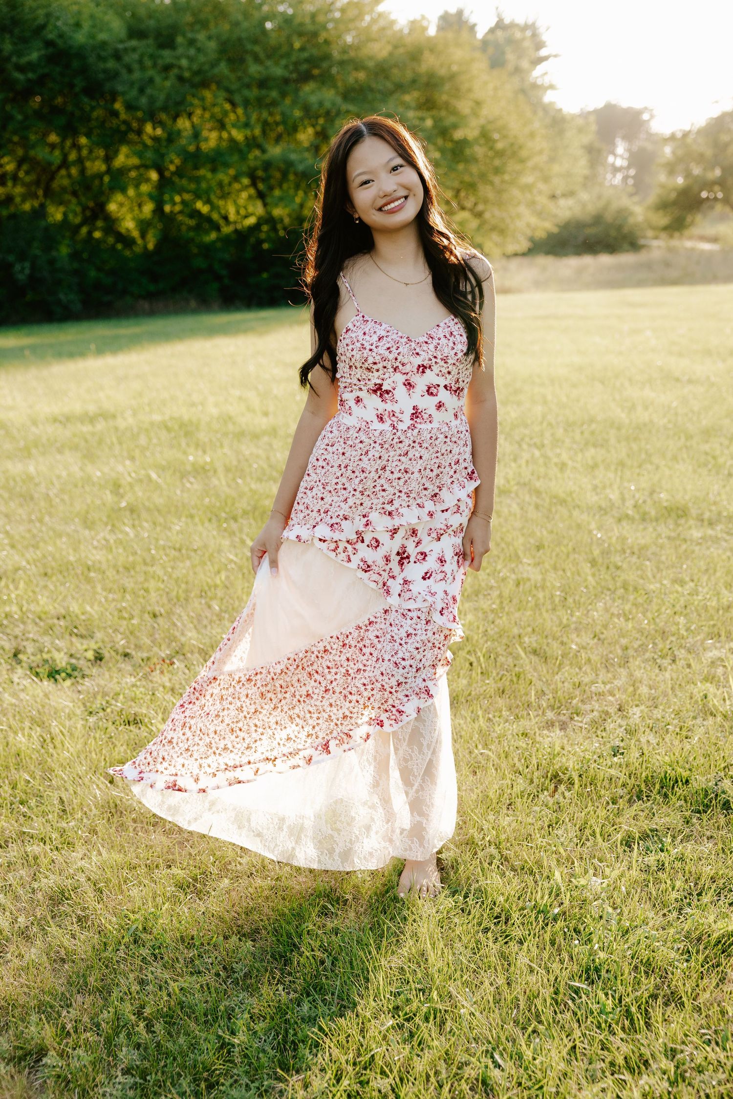 A girl in a white floral dress twirls playfully in a sunlit meadow surrounded by green trees at Pioneers Park in Lincoln Nebraska.