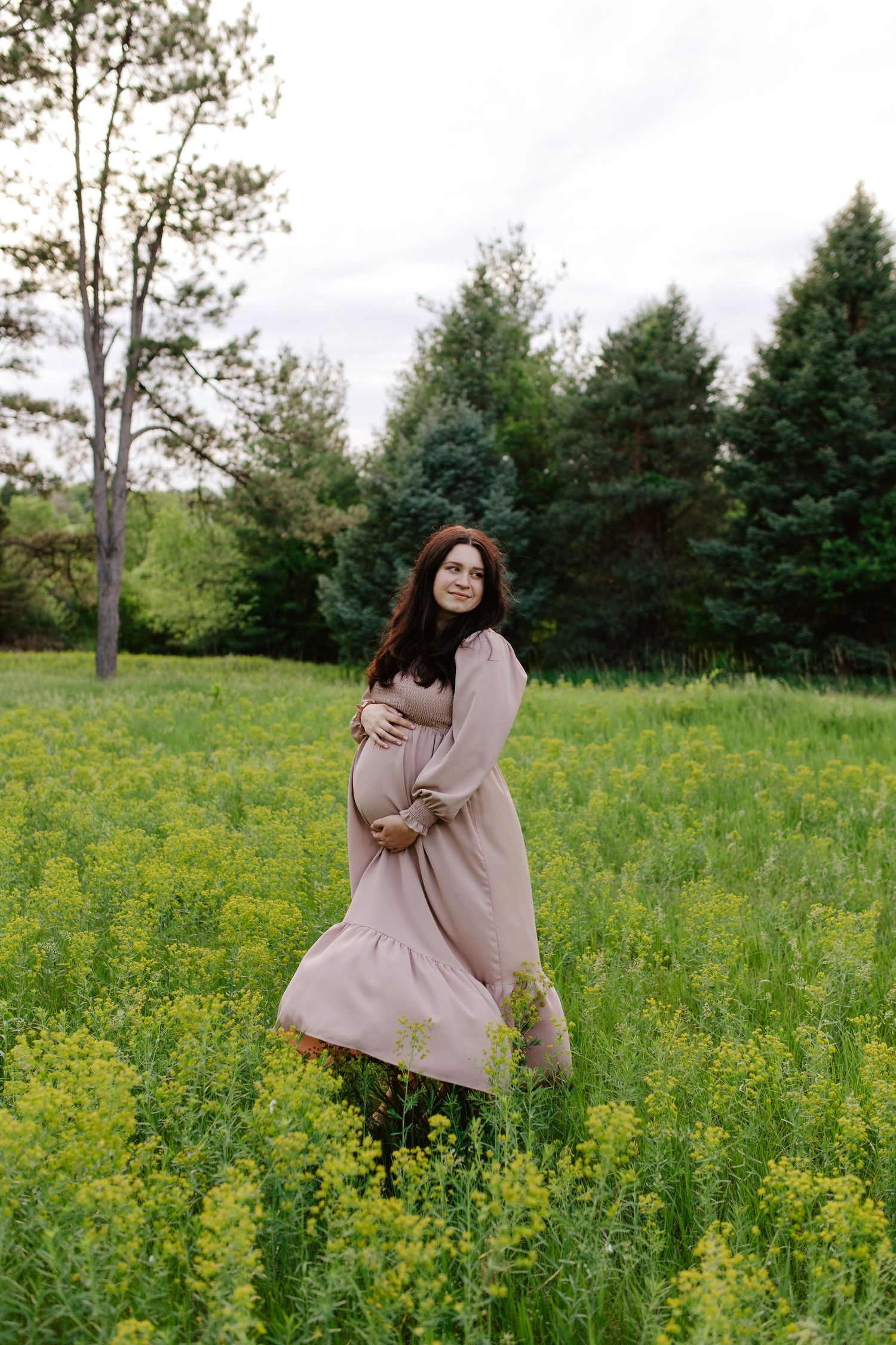 A woman in a flowing blush dress sits peacefully in a wildflower meadow with evergreen trees in the background.