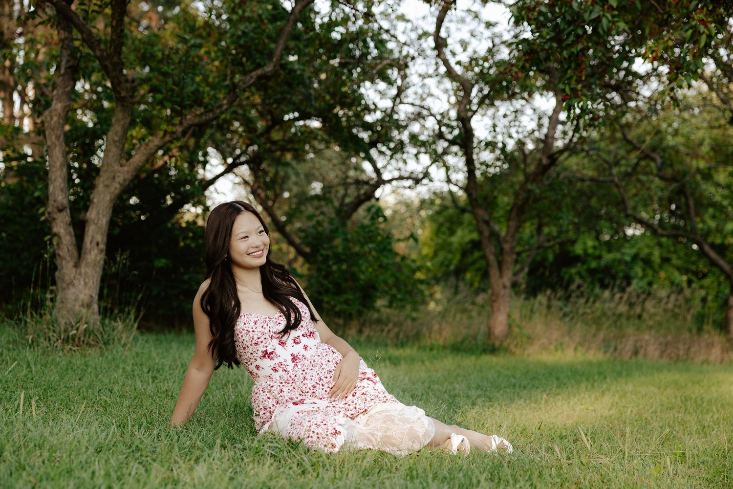A girl in a red and white floral dress sits on green grass during sunset for her senior photo session.