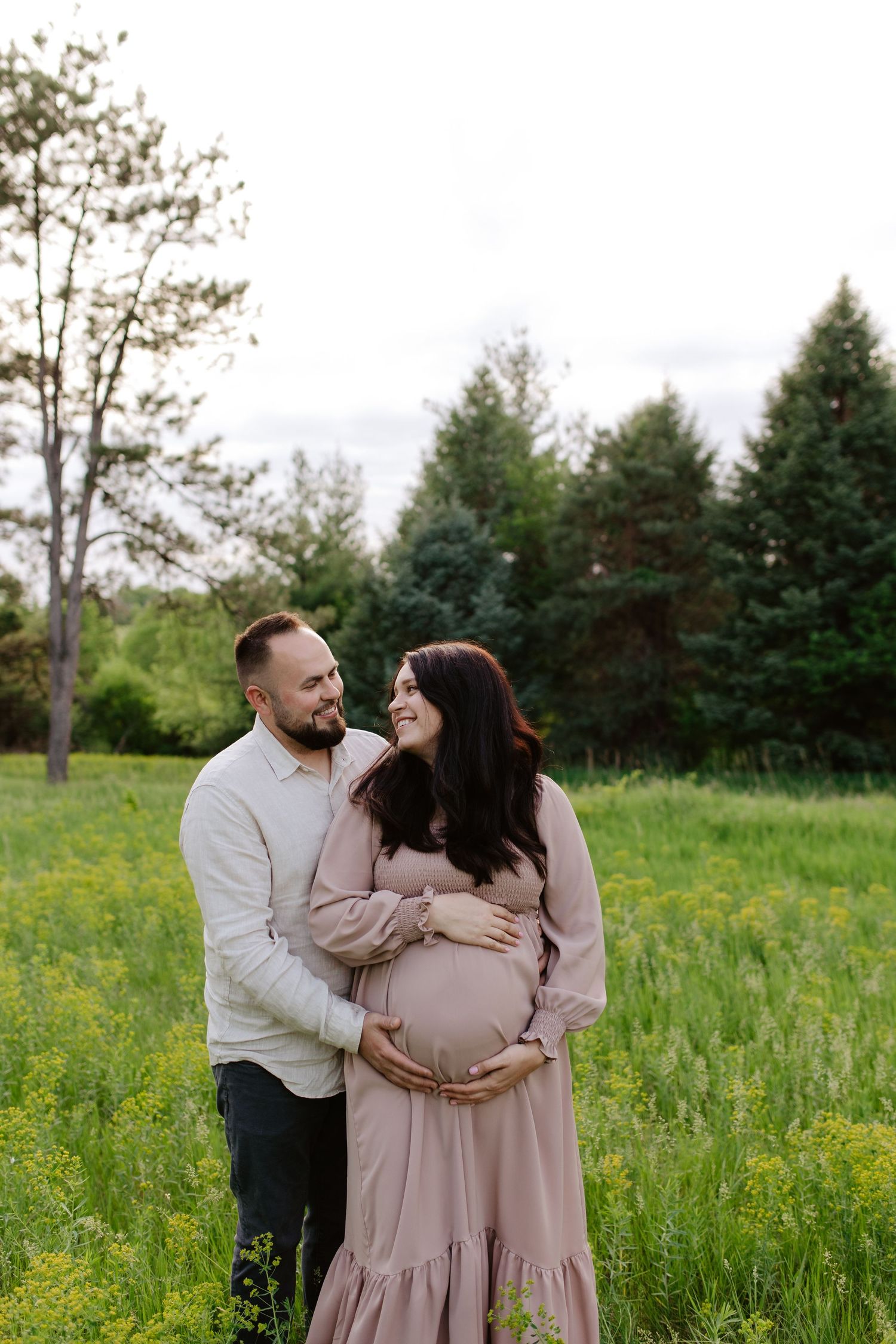 Expectant couple embraces in a spring meadow with yellow wildflowers and evergreen trees in the background.