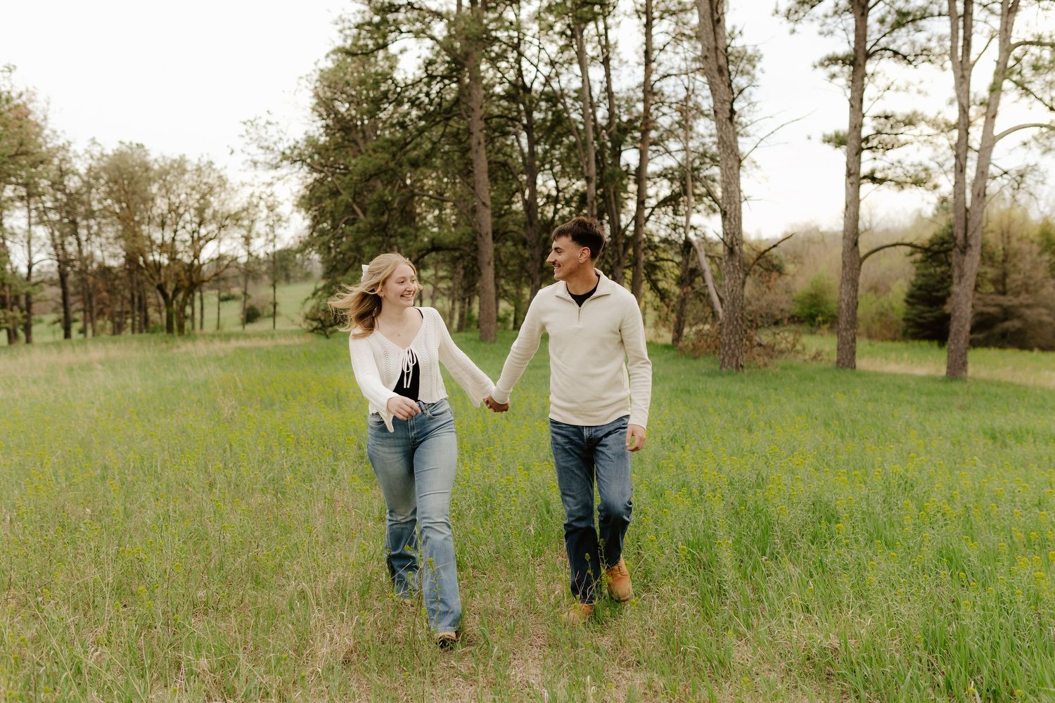 A couple walks hand in hand through a sunny green meadow with tall trees in the background.