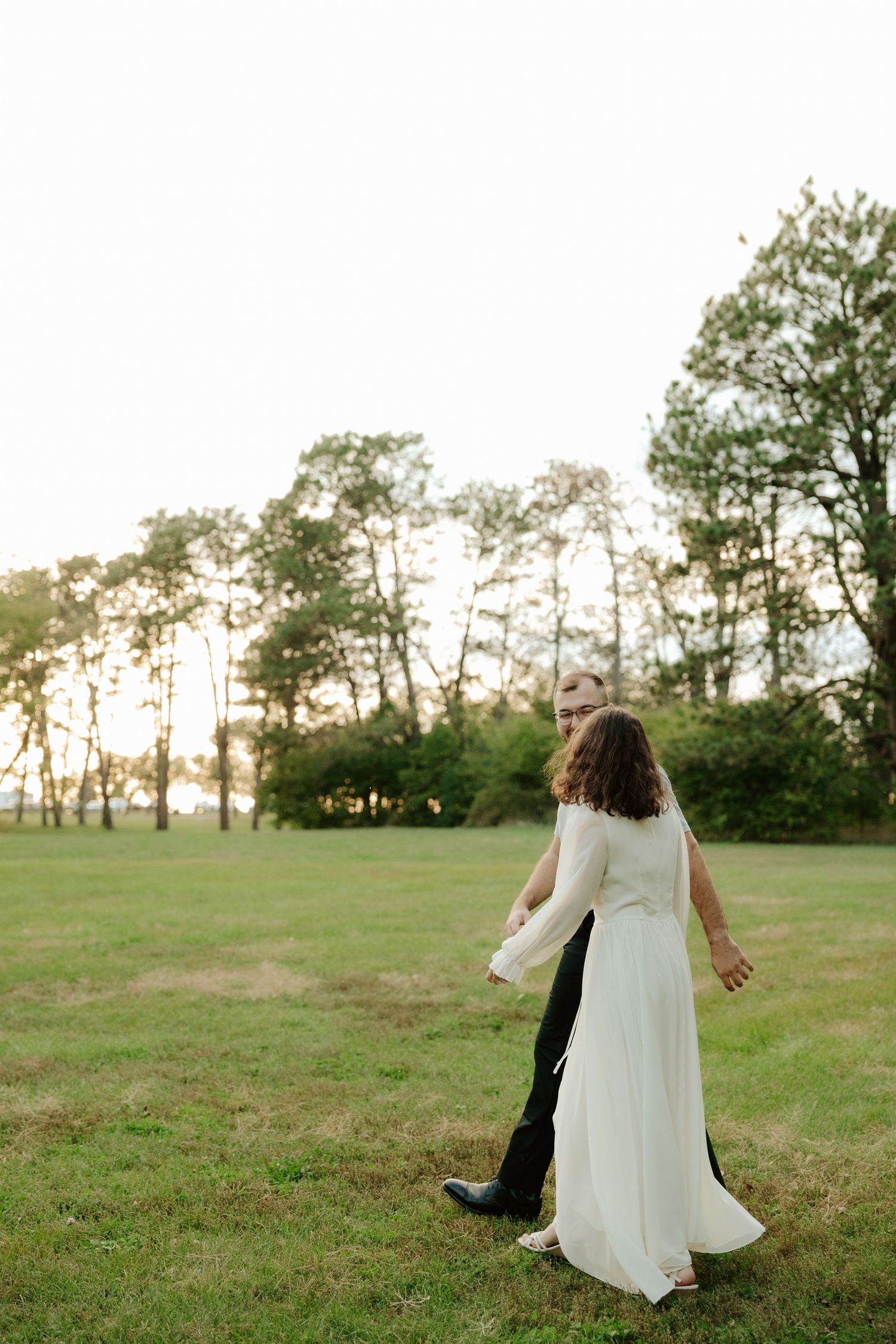 A couple dances together in a grassy field with autumn trees in the background during a romantic outdoor moment.