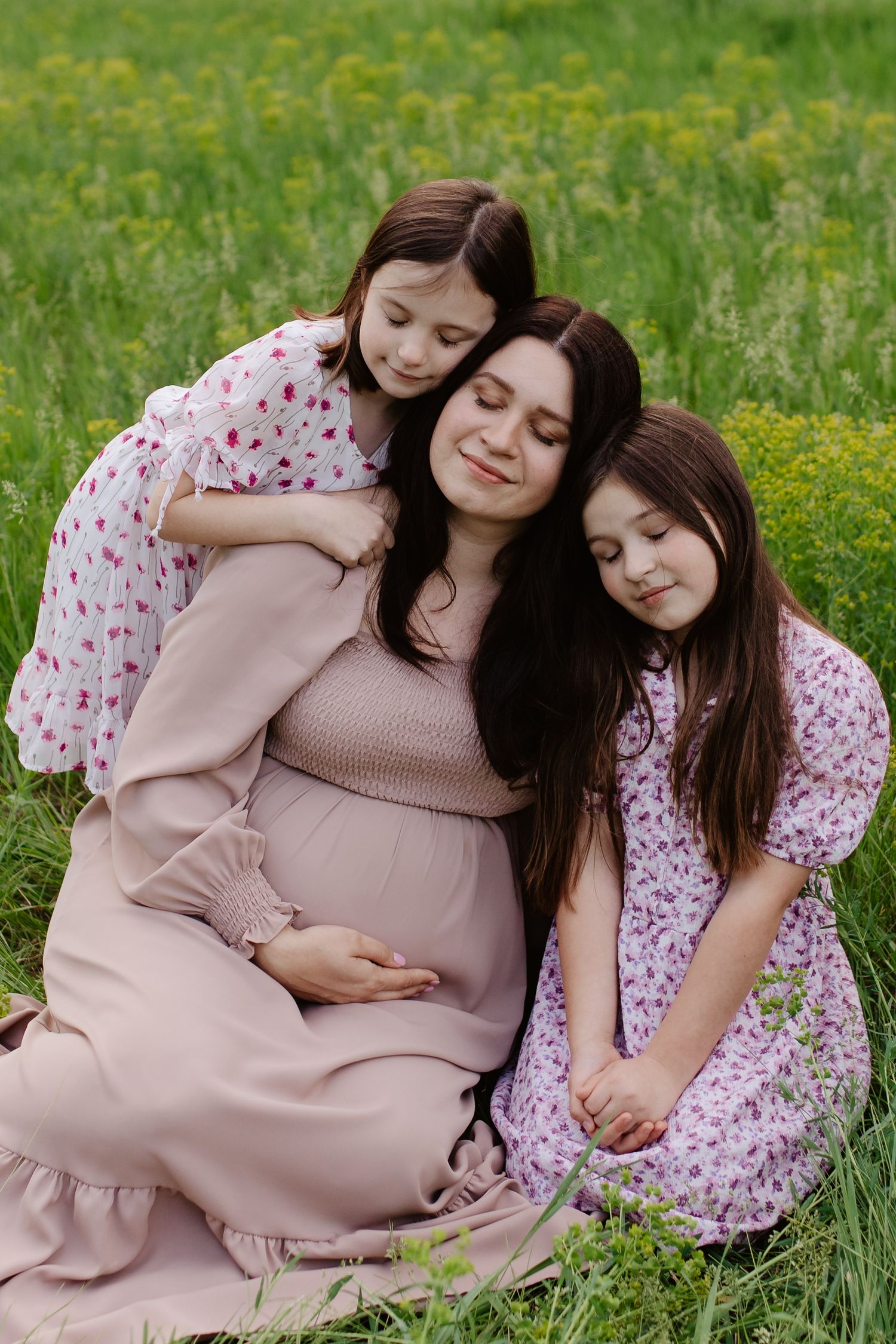 Mother and daughters in pink dresses sit together on grass with yellow wildflowers enjoying a tender moment.