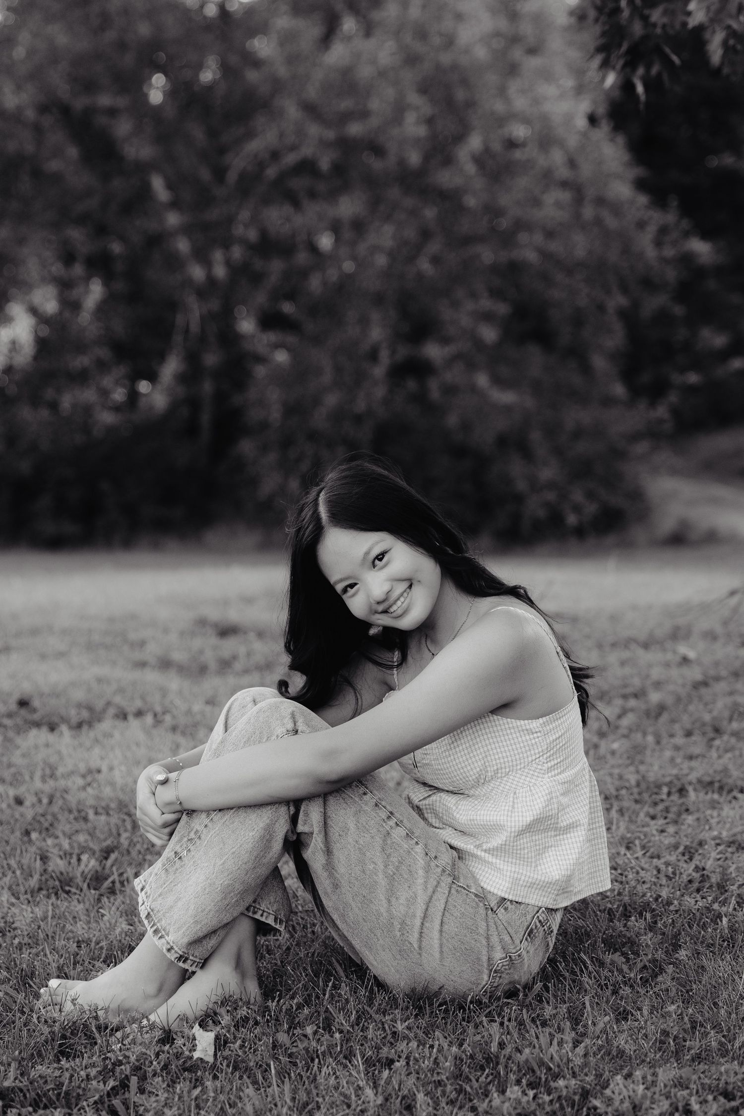A girl in a beige top and light jeans sits on green grass with trees in the background during golden hour at Pioneers Park in Lincoln NE.