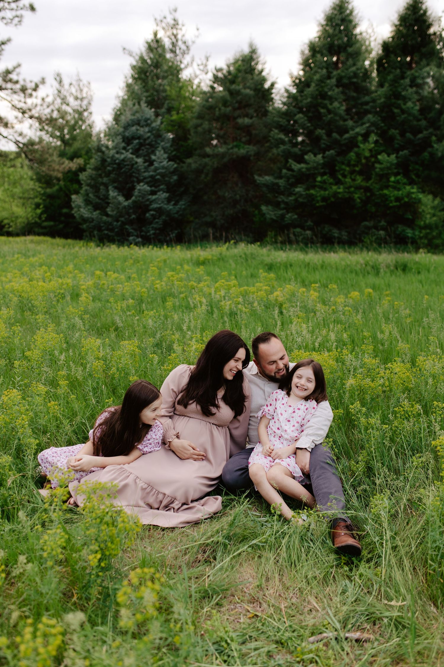 A family sits together in a green meadow with wildflowers and evergreen trees in the background.