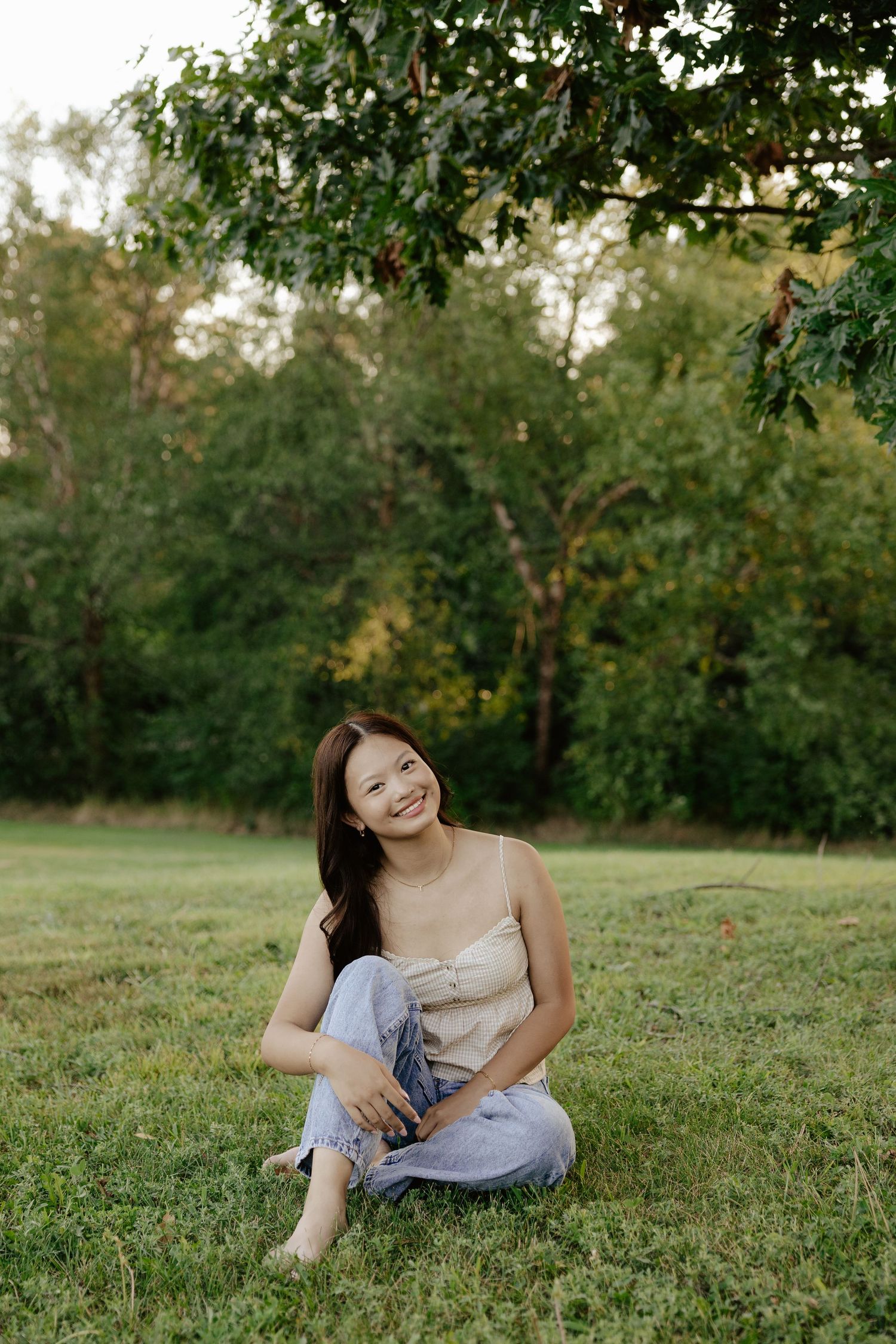 Girl sitting on grass in a casual outfit with trees in background during golden hour lighting, posing for senior photos.