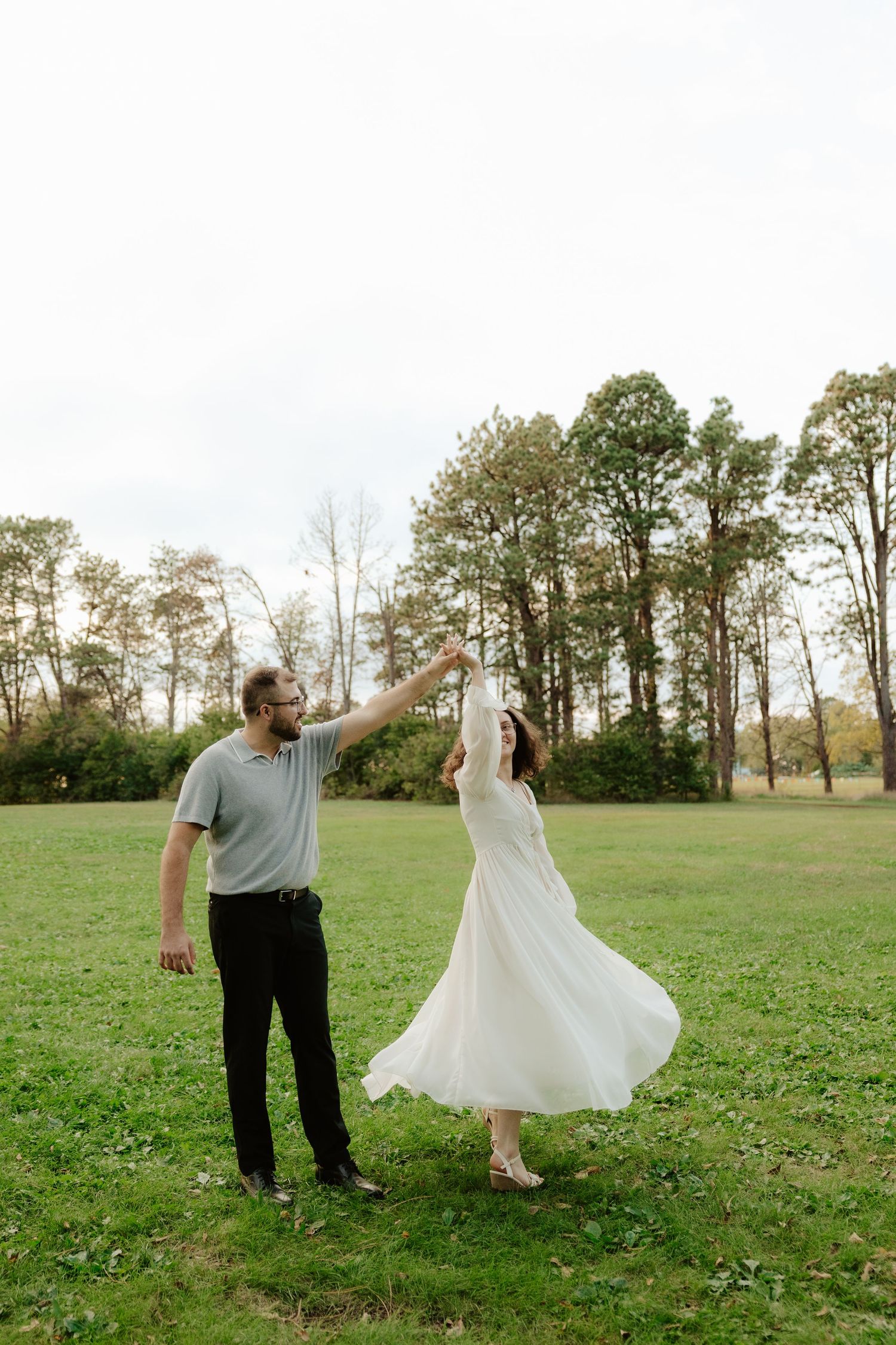 A couple dances together in a grassy field with autumn trees in the background during a romantic outdoor moment.