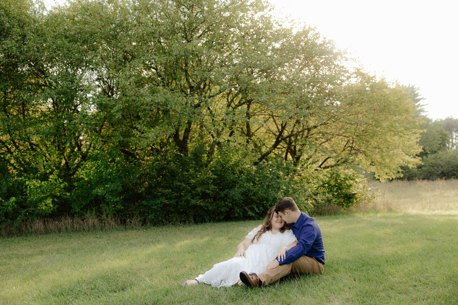 A couple sits together on grass in a natural outdoor setting surrounded by green trees and foliage at Pioneers Park in Lincoln Nebraska.