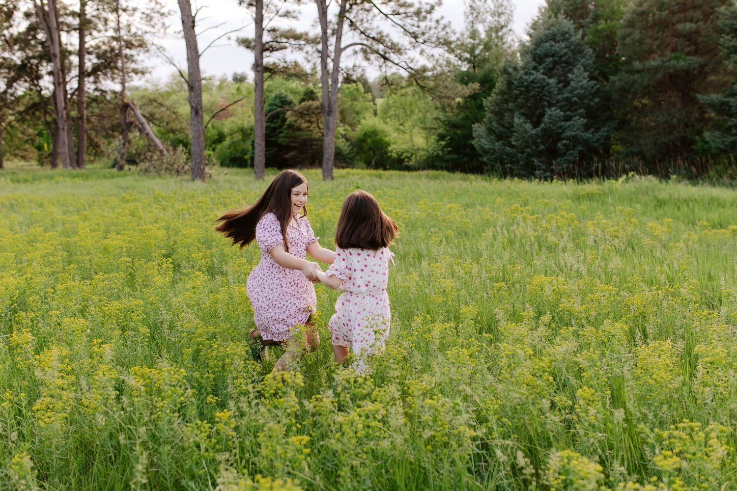 Two sisters in flowy dresses run together through a sunlit meadow filled with yellow wildflowers.