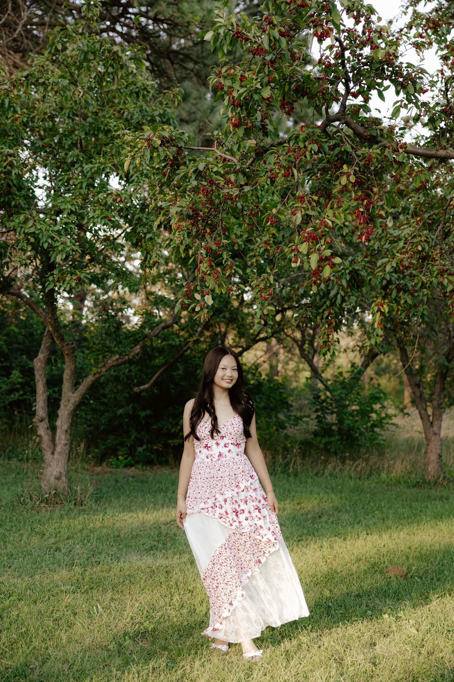 A girl in a white and red floral dress poses outdoors under blooming trees on a sunny day for her senior photos.