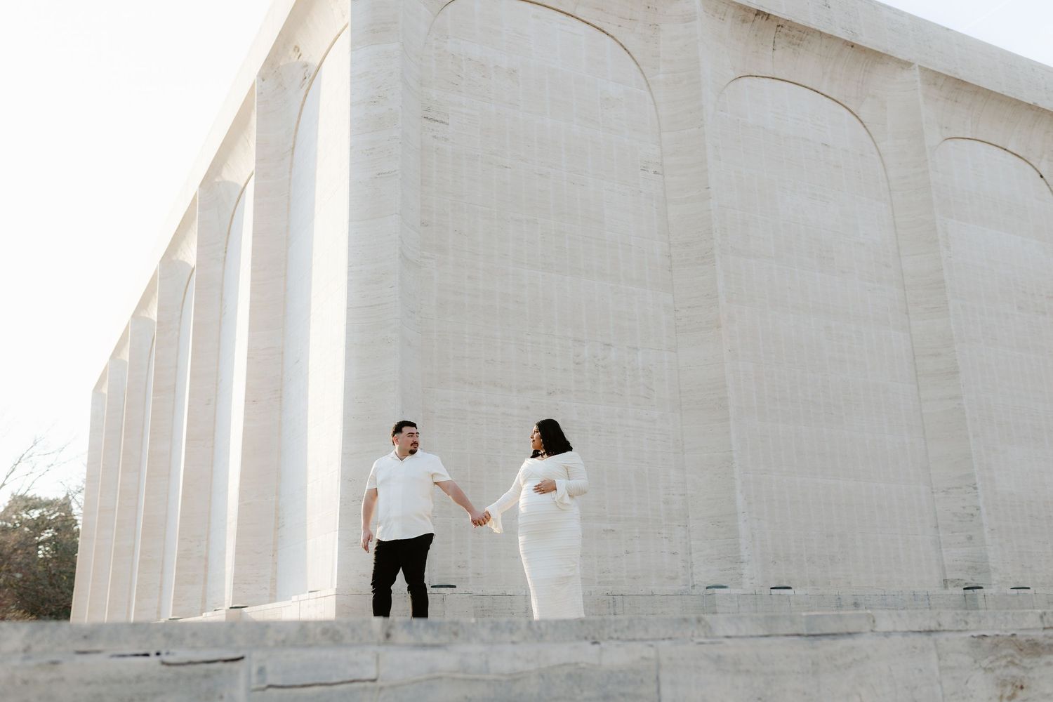 Couple holding hands while standing against white arched concrete architectural structure during a maternity photoshoot.