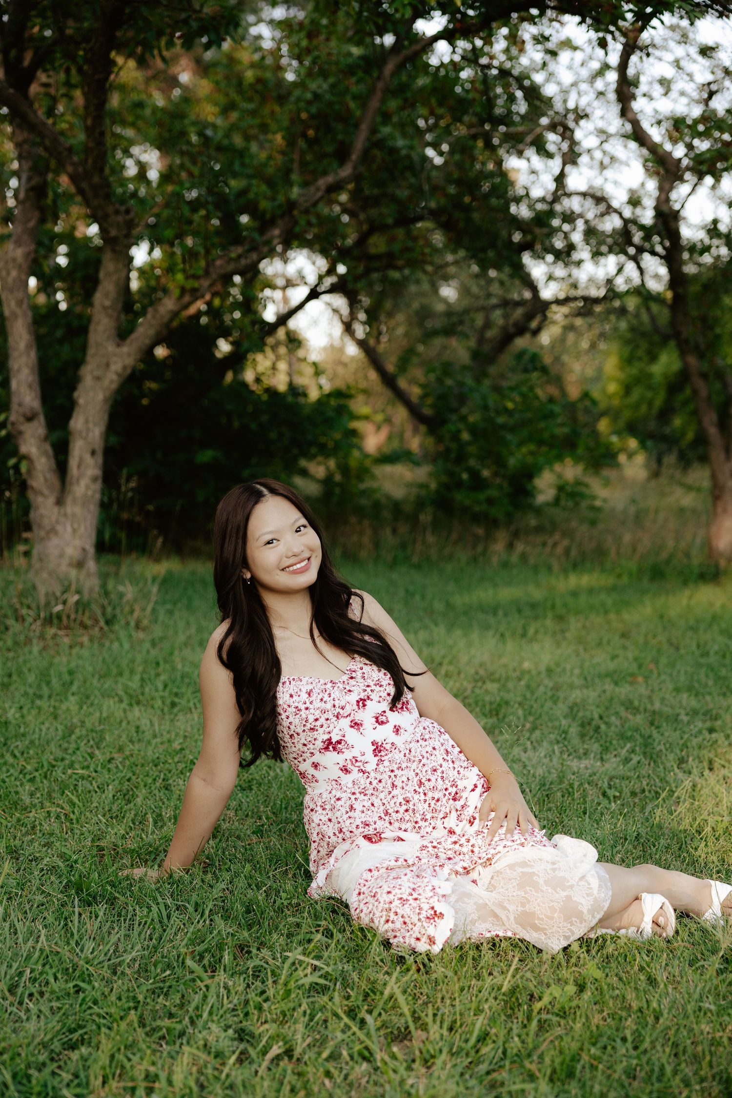 A girl in a red and white floral dress sits on green grass during sunset for her senior photo session.