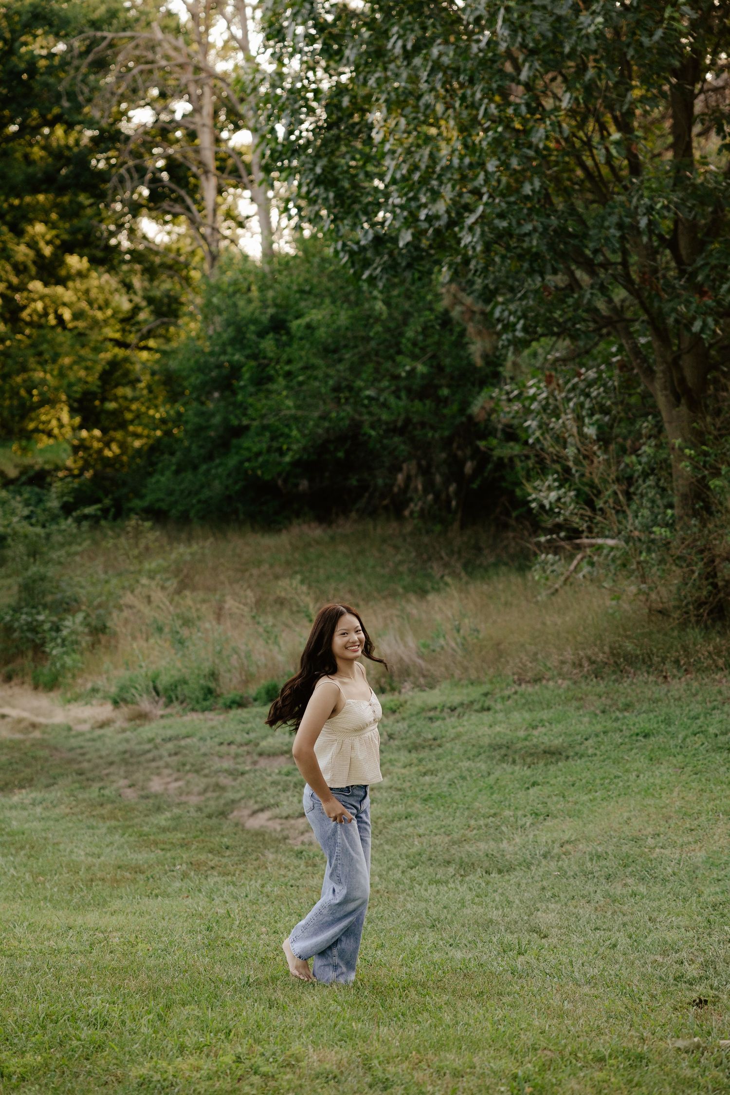 A girl walking through a grassy field in casual clothing at sunset for her senior photo session.