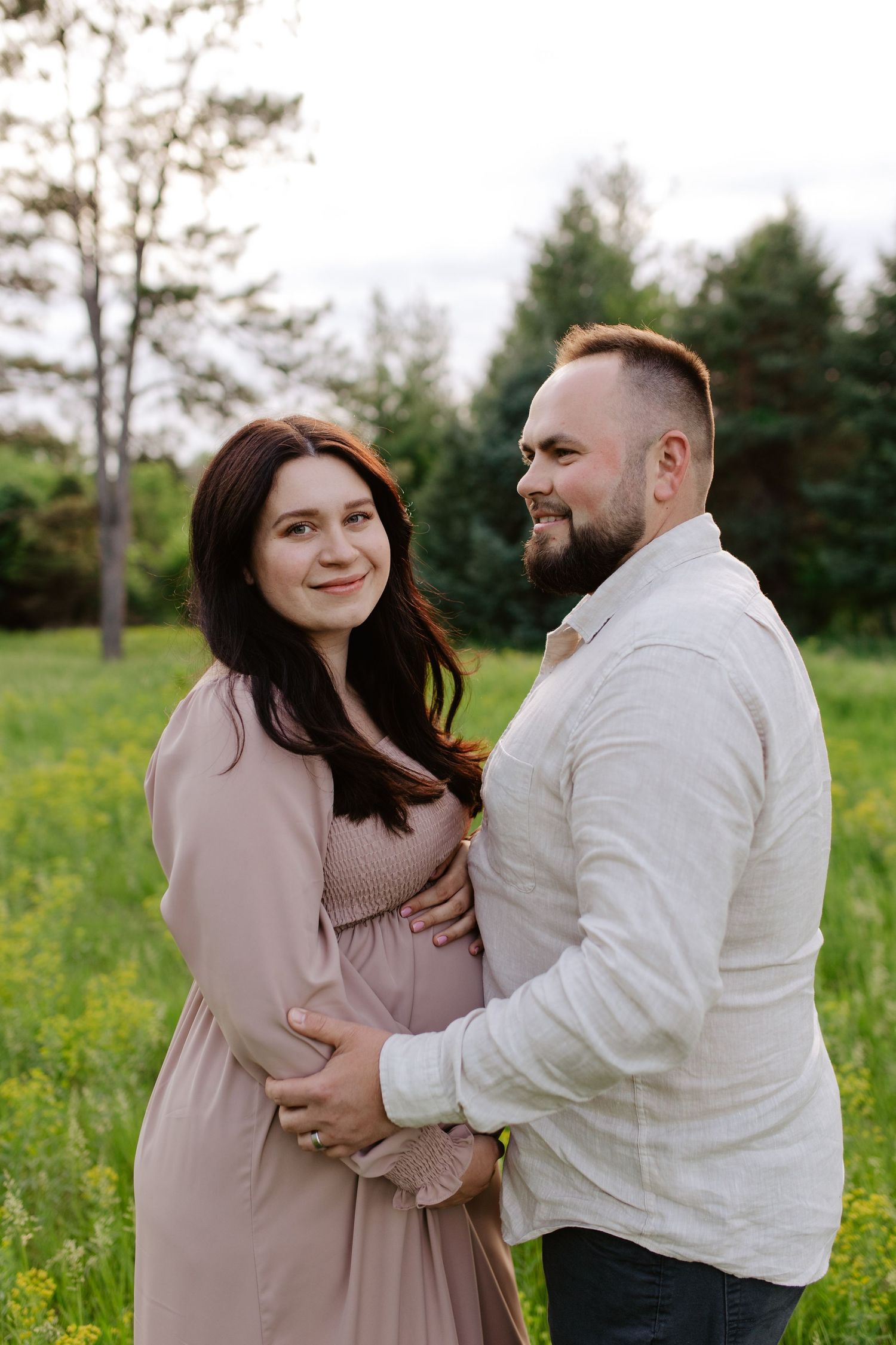 A couple shares a tender moment in a grassy field with pine trees, as they embrace during a maternity photoshoot.
