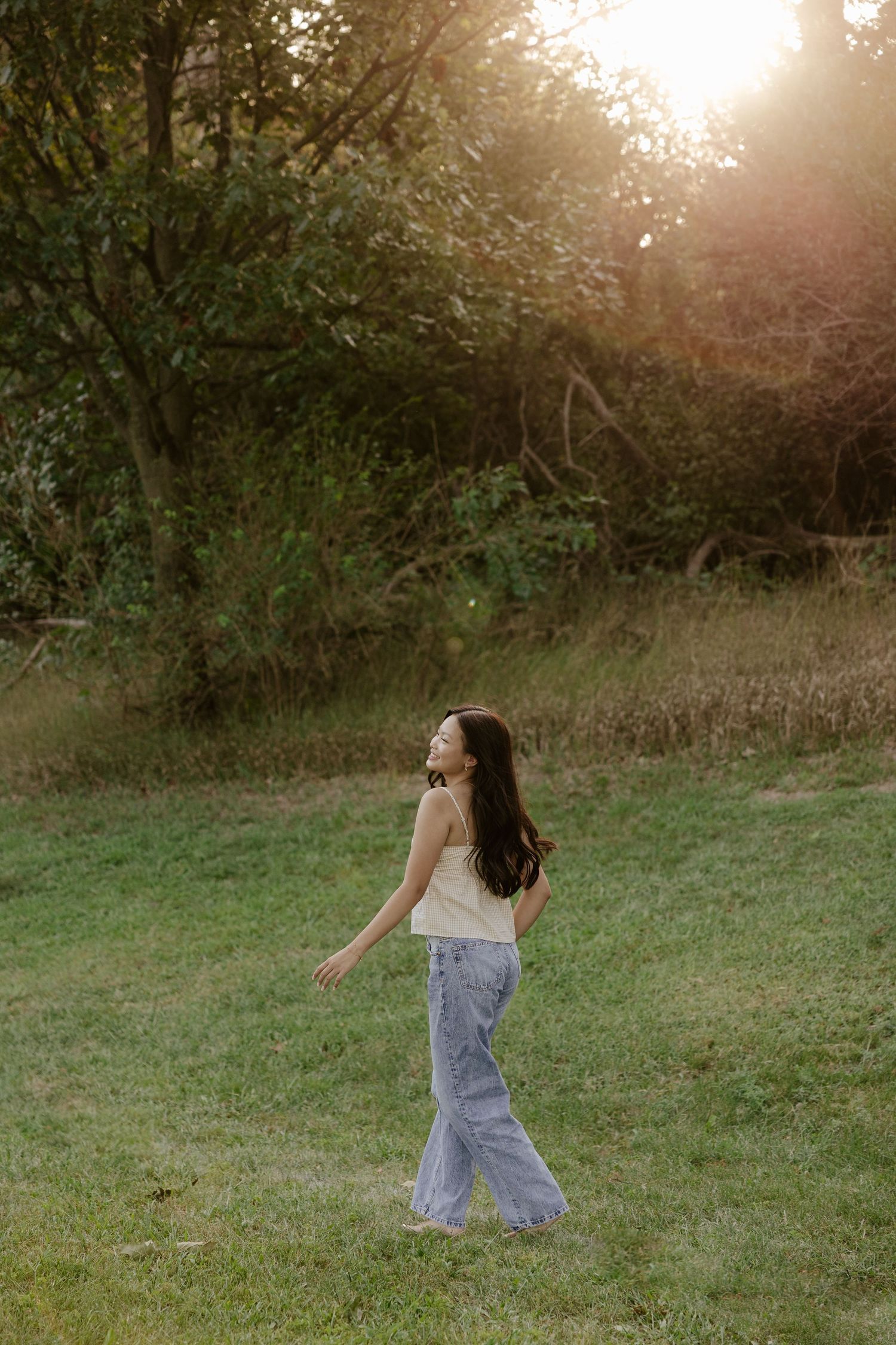 A girl walking through a grassy field in casual clothing at sunset for her senior photo session.