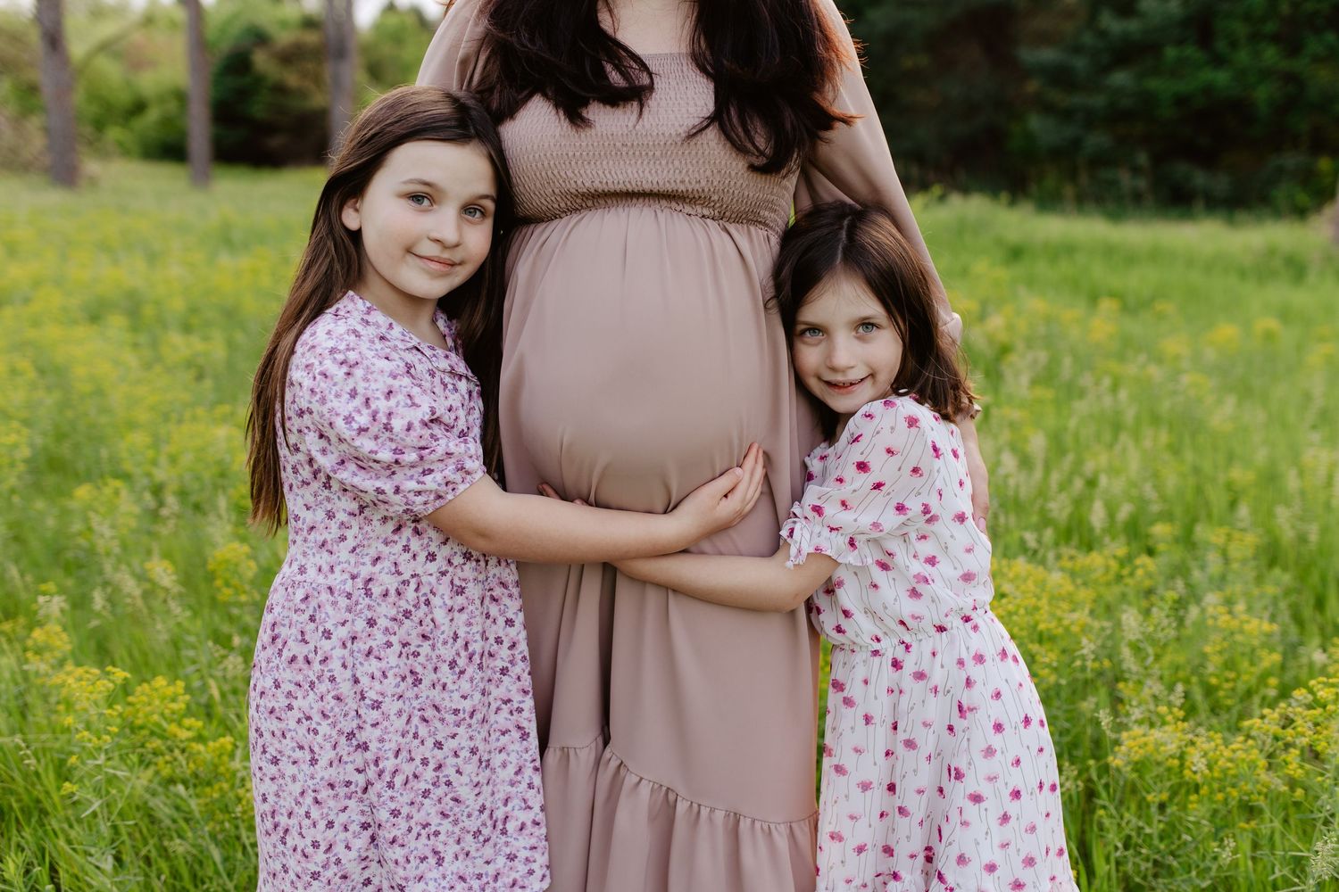 Mother and daughters share a tender moment in a grassy field with pine trees, as they embrace during a maternity photoshoot.