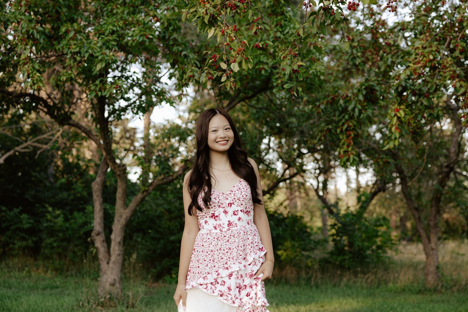 A girl in a white and red floral dress poses outdoors under blooming trees on a sunny day for her senior photos.