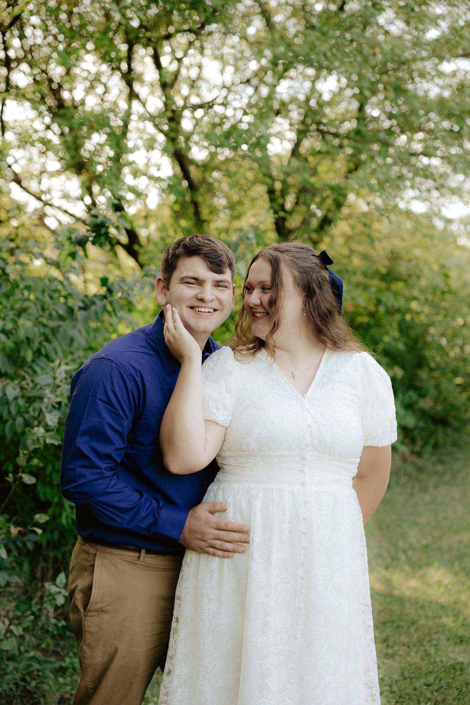 A couple poses together outdoors in wedding attire with white dress and navy blue shirt among green foliage.
