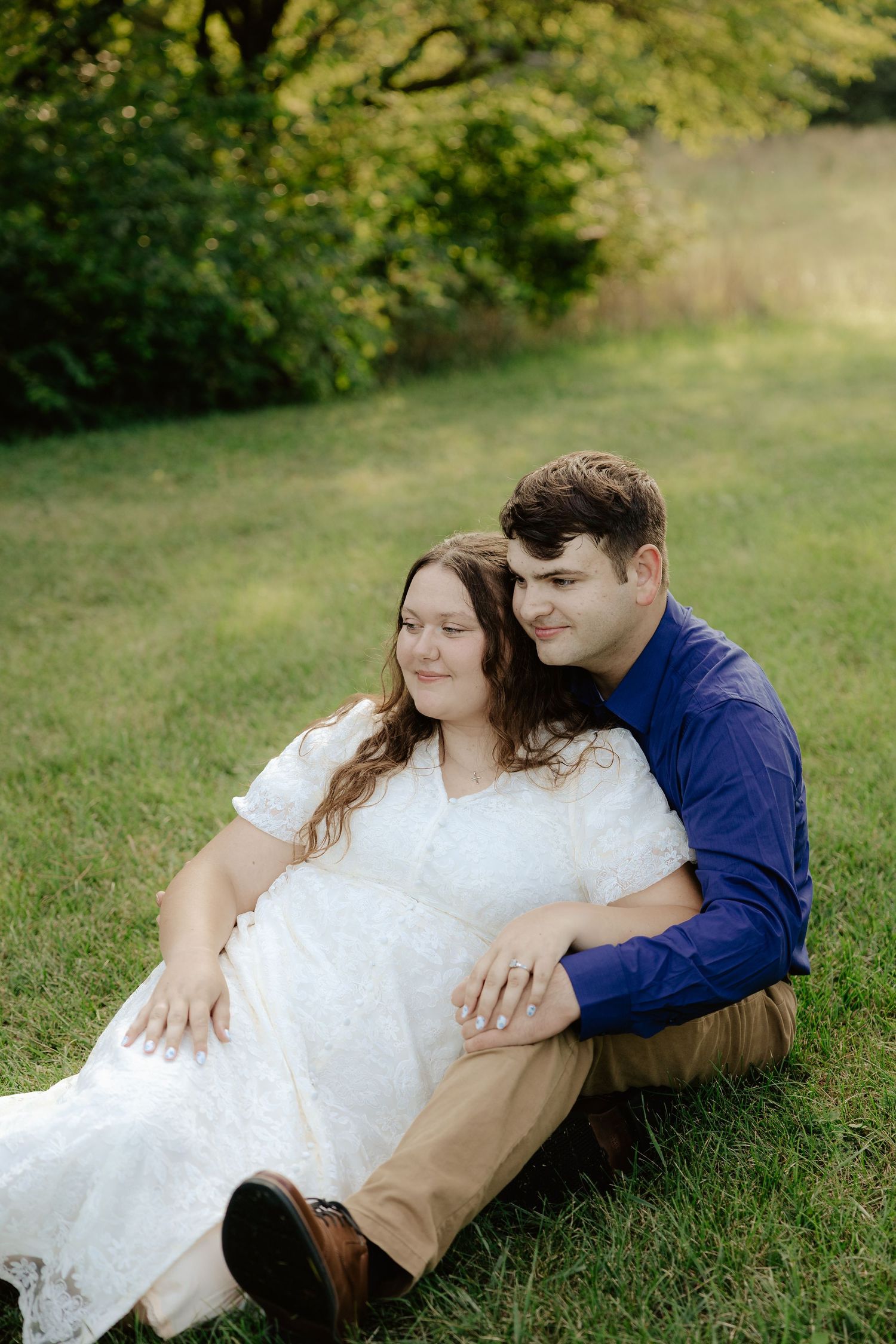 A couple sits together on grass in a natural outdoor setting surrounded by green trees and foliage at Pioneers Park in Lincoln Nebraska.